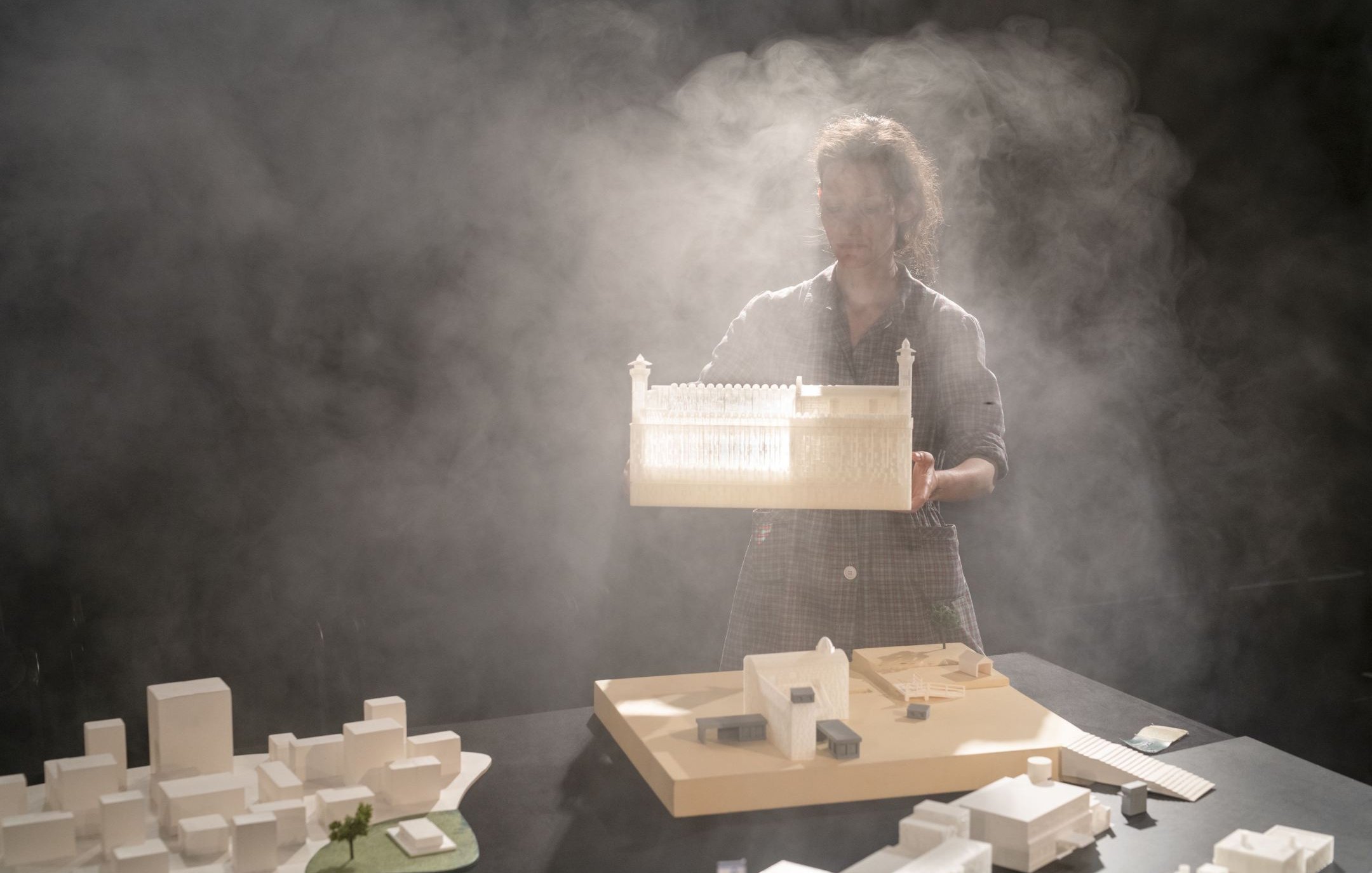L'image montre une personne tenant une maquette d'un bâtiment, dans un environnement brumeux. Le fond est sombre, ce qui met en valeur la maquette et la silhouette de la personne. Sur la table, il y a d'autres maquettes et éléments architecturaux, suggérant une scène de travail ou de présentation liée à l'architecture. La lumière semble filtrer à travers la brume, créant une ambiance mystérieuse et artistique.