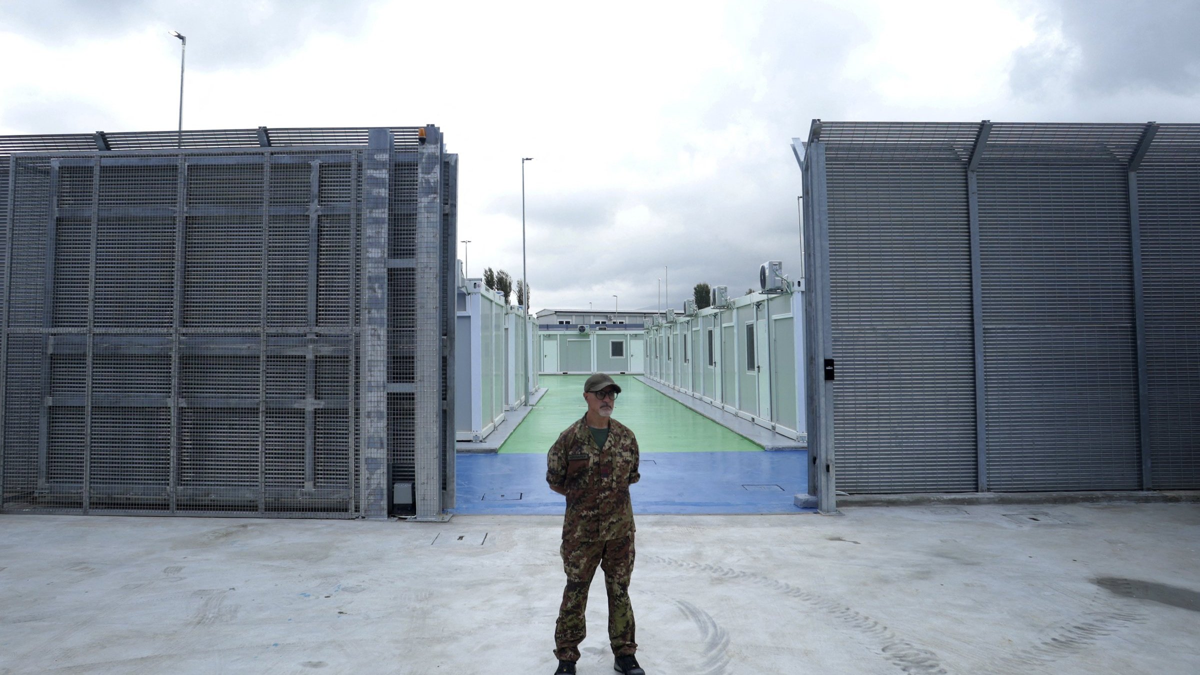 L'image montre un soldat en uniforme qui se tient au centre d'une zone sécurisée, probablement à l'extérieur d'une installation. À l'arrière-plan, on peut voir des structures métalliques grises et des murs, créant une ambiance de confinement. Le sol est en béton, et il y a une passerelle colorée qui semble être verte ou bleue. Le ciel est nuageux, suggérant un temps sombre. L'ensemble de l'image évoque un sentiment de sécurité renforcée et de surveillance.