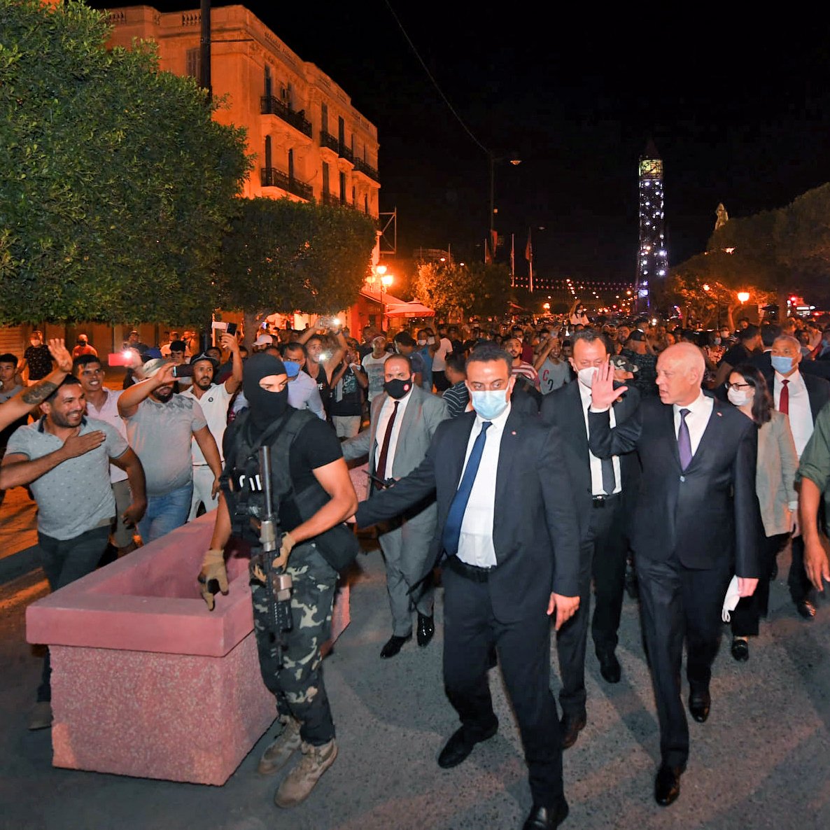 The image depicts a nighttime scene in a city, where a group of people is gathered in a street. In the foreground, a group of officials, including one dressed in a suit, is walking through a crowd, accompanied by a person in tactical gear, suggesting a security presence. The atmosphere appears charged, with some people in the crowd gesturing expressively. Trees and buildings with lights are visible in the background, contributing to the nighttime setting. Overall, the scene conveys a sense of political or social gathering, possibly in response to current events.