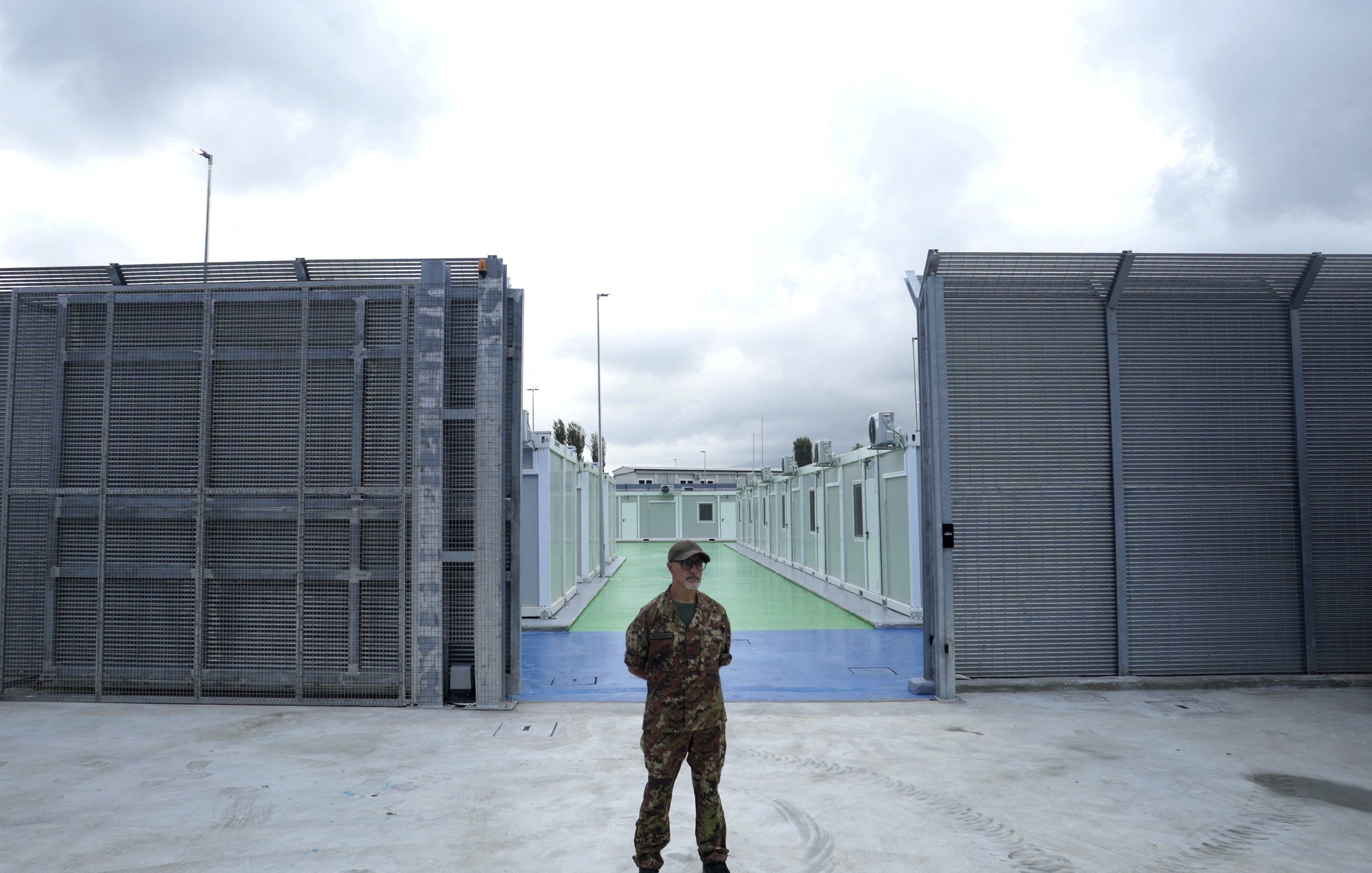 L'immagine mostra un soldato in uniforme, in piedi al centro di un'area delimitata. Sullo sfondo si possono vedere due strutture metalliche grigie che sembrano essere recinzioni, con un'area di pavimentazione colorata di verde e blu tra di esse. Il cielo è nuvoloso, suggerendo una giornata grigia. L'atmosfera sembra essere di sicurezza e controllo, con un forte senso di vigilanza da parte del soldato.
