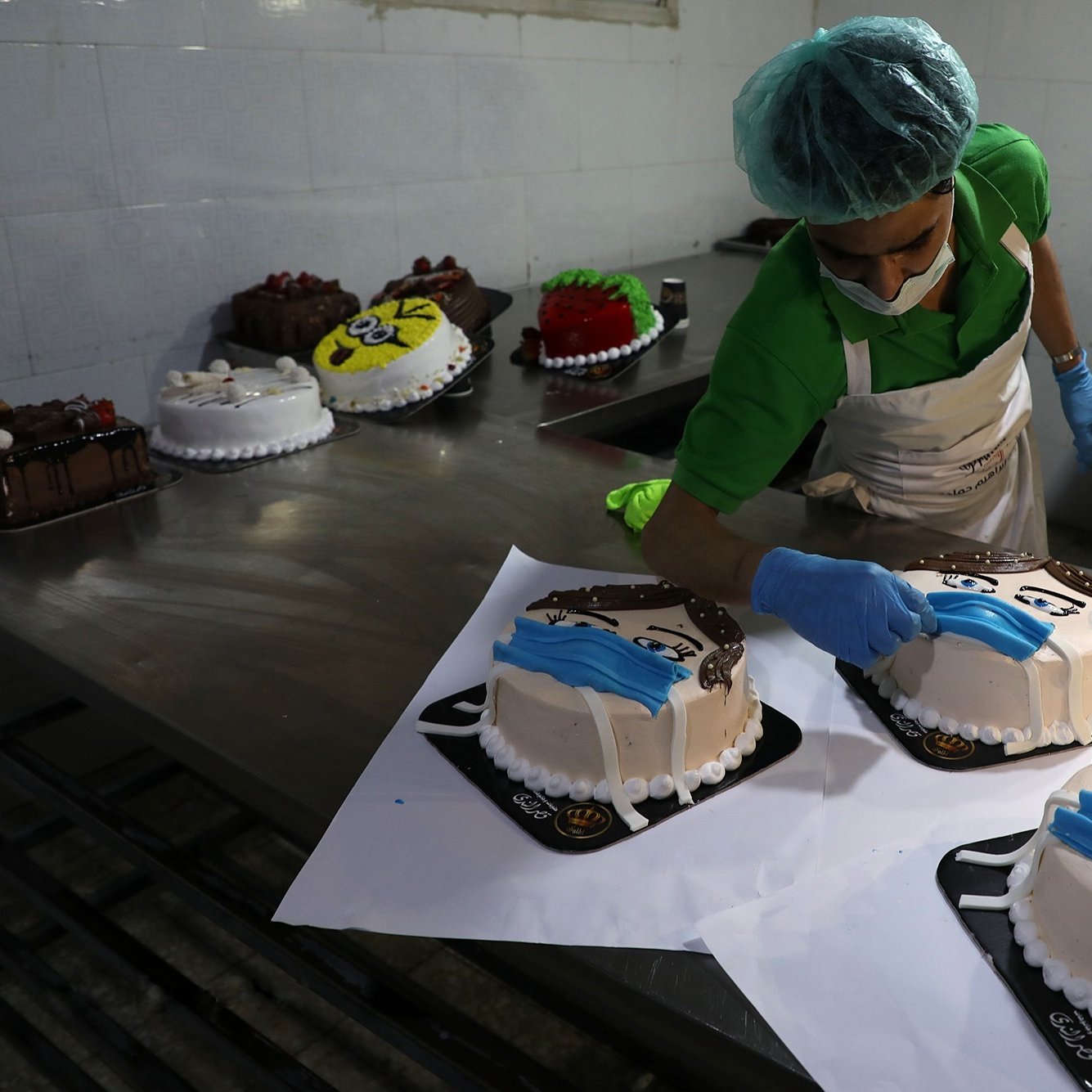 در تصویر یک نانوایی یا قنادی دیده می‌شود که یک کارگر در حال تزئین کیک‌هاست. این کیک‌ها شکل و طراحی خاصی دارند و بر روی آن‌ها معمولاً تصاویر چهره‌ای با ماسک قرار داده شده است. کارگر با دقت کیک‌ها را تزئین می‌کند و از دستکش و کلاه مخصوص استفاده می‌کند. در پس‌زمینه، تعدادی کیک دیگر نیز قرار دارند که آماده فروش هستند. فضای قنادی تمیز و مرتب به نظر می‌رسد.