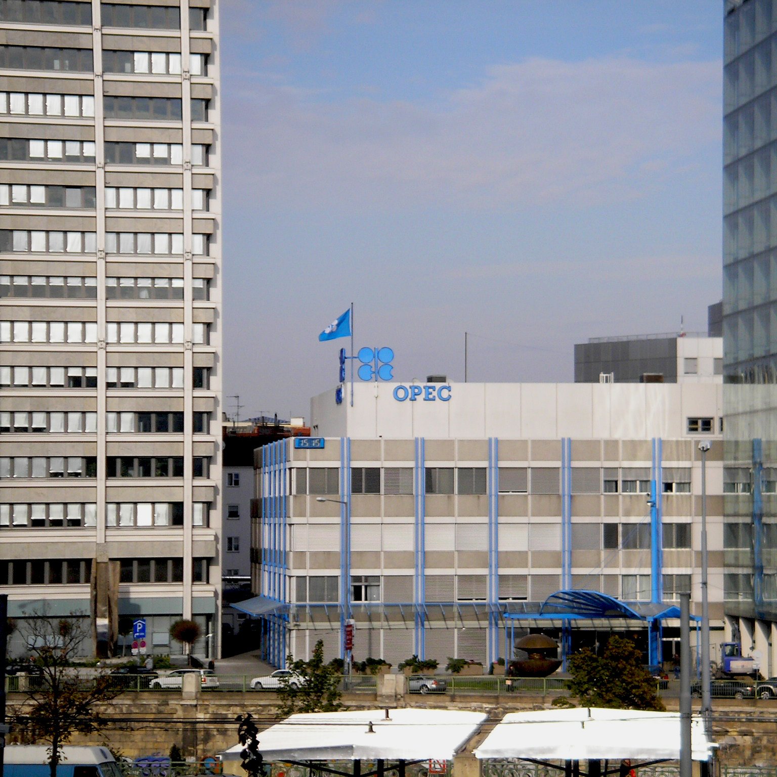 الصورة تظهر مبنى منظمة الدول المصدرة للنفط (أوبك) في وسط مدينة حديثة. يتكون المبنى من عدة طوابق ويتميز بتصميمه العصري. يوجد علم أوبك يرفرف فوق المبنى، كما أن هناك نوافذ كبيرة تسمح بدخول الضوء الطبيعي. المحيط يتضمن مبانٍ عالية أخرى، مما يشير إلى منطقة حضرية نشطة. الأجواء تبدو واضحة مع سماء زرقاء.