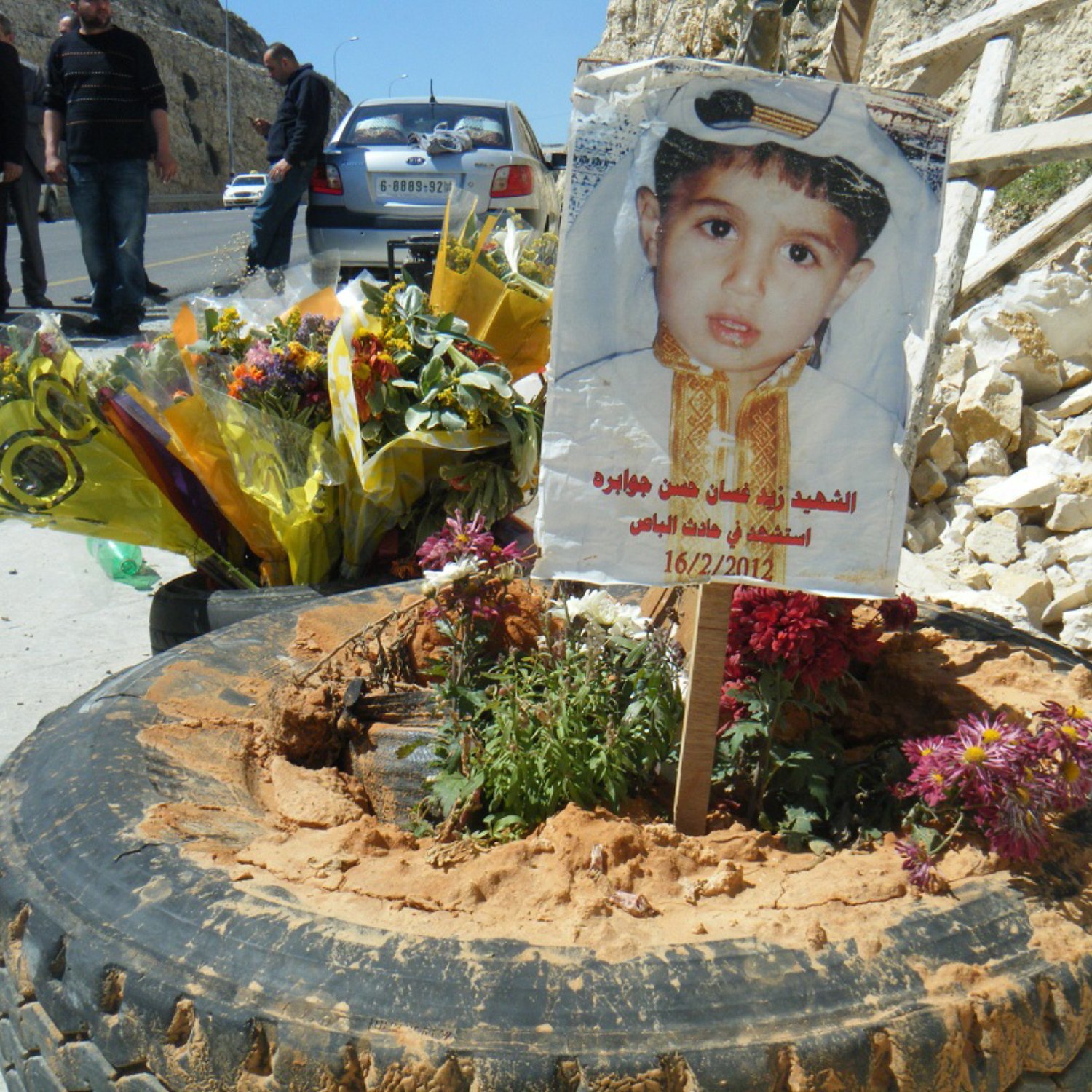 L'immagine mostra un tributo commemorativo collocato su un terreno. Al centro, c'è un cartello che presenta un ritratto di un bambino, adornato con un abbigliamento tradizionale. Attorno al cartello ci sono fiori freschi e piante, posizionati in una gomma di automobile. Il tutto è circondato da un ambiente duro, con rocce visibili e alcune persone in fondo che sembrano partecipare a una commemorazione. La data sul cartello indica un giorno significativo, possivelmente legato alla memoria del bambino.
