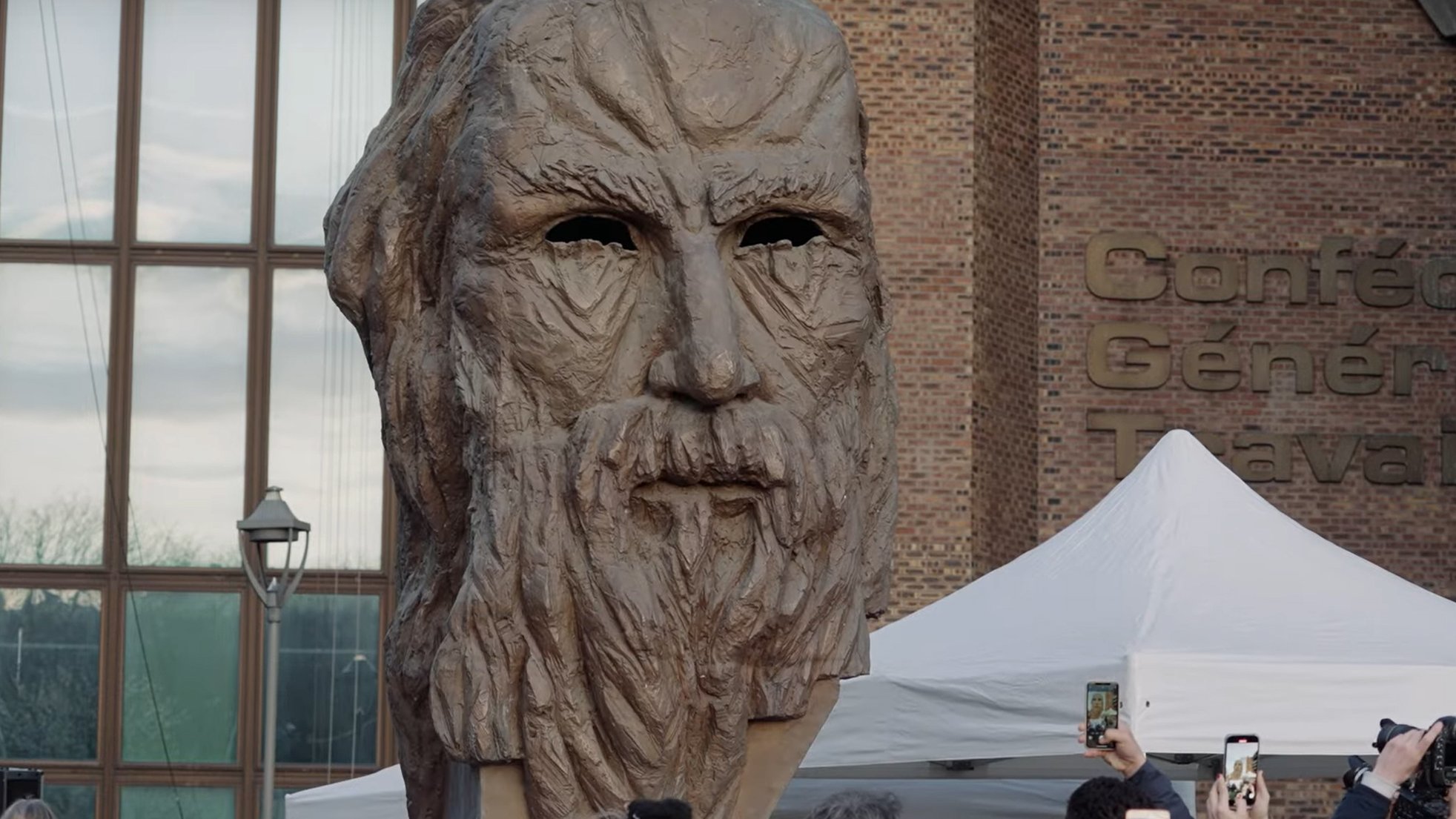L'image montre une grande sculpture représentant un visage ancien, probablement celui d'une figure historique ou mythologique, avec des traits marqués et une barbe. Elle est entourée d'une foule de personnes qui semblent prendre des photos avec leurs téléphones. Il y a aussi des tentes et un bâtiment visible en arrière-plan, ce qui indique qu'il s'agit probablement d'un événement public ou d'une exposition. L'atmosphère semble être festive et attentive, centrée sur l'œuvre sculpturale.