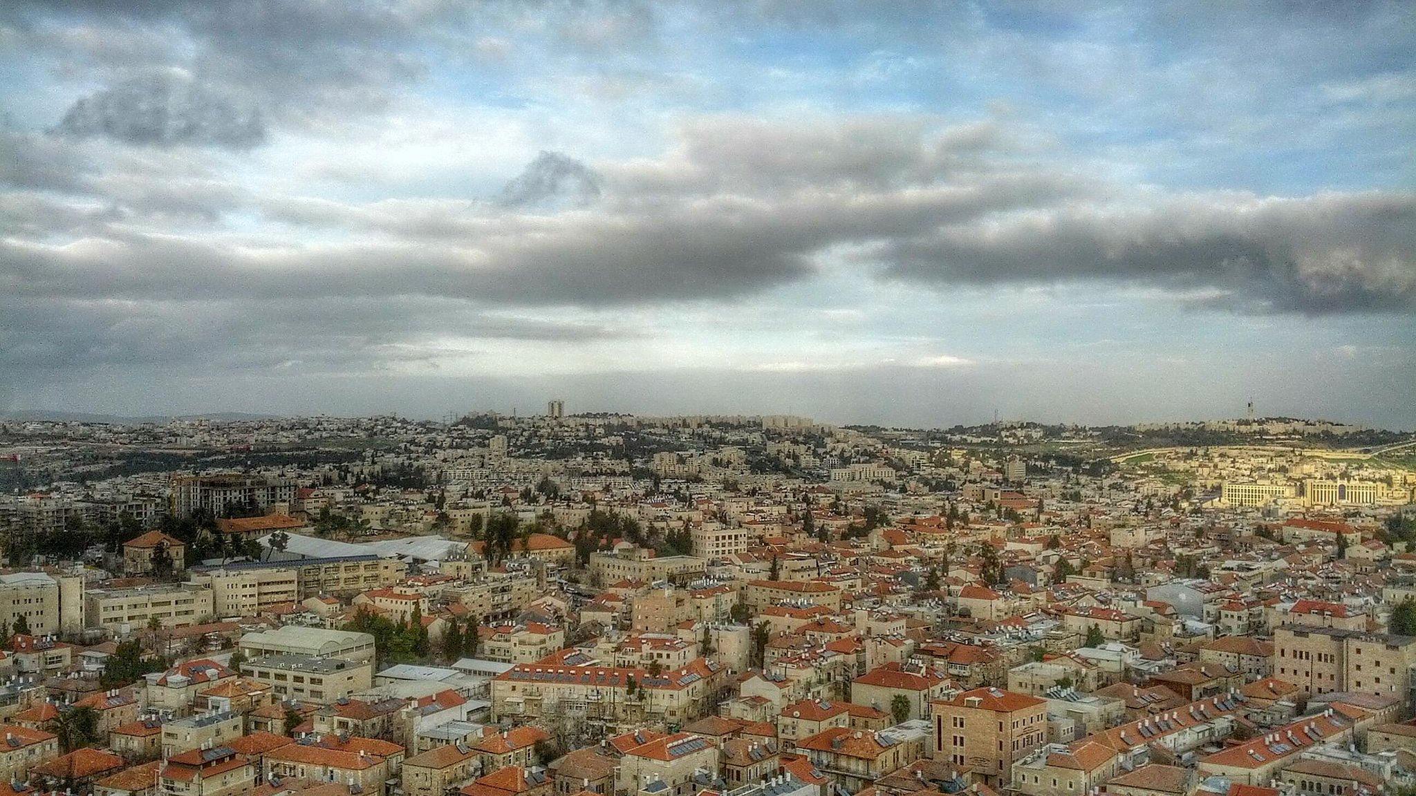 L'image montre une vue panoramique d'une ville, probablement Jérusalem, avec des bâtiments disposés sur plusieurs collines. Les toits sont principalement en tuiles rouges, et il y a de nombreux édifices en pierre. Le ciel est nuageux, ajoutant à l'ambiance de la scène, avec une lumière douce qui éclaire la ville. On peut également apercevoir des zones de verdure entre les bâtiments, ce qui ajoute de la profondeur au paysage urbain.