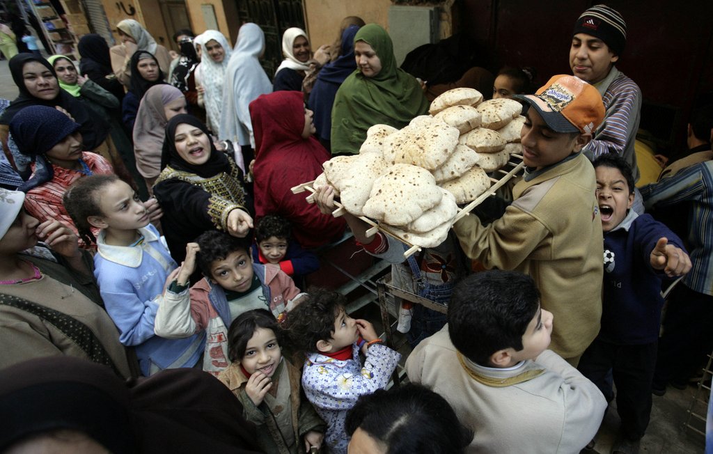 L'image montre une scène animée dans une rue, probablement dans un pays arabe. Un jeune garçon porte un plateau chargé de pains, entouré d'une foule de personnes, comprenant des adultes et des enfants. On peut voir des visages souriants et des expressions variées, ce qui donne une impression de convivialité et d'effervescence. Les gens semblent attendre pour acheter du pain, et la diversité des vêtements suggère une ambiance culturelle riche. L'image capte un moment de vie quotidienne, centré sur l'échange et la nourriture.
