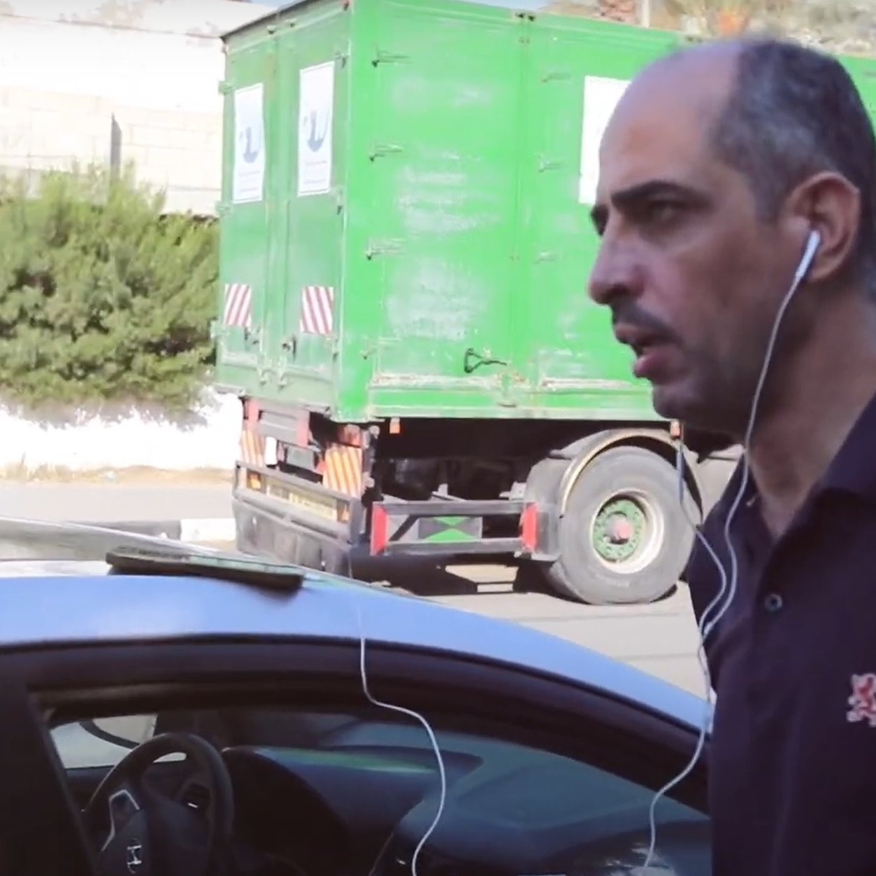 Nell'immagine si vede un uomo che cammina accanto a un'auto. Indossa una maglietta scura e ha delle cuffie che gli pendono dalle orecchie. Sullo sfondo si nota un camion verde e alcune piante. L'uomo sembra concentrato e diretto verso un punto specifico. A sinistra, è presente un'etichetta che indica il suo nome e la sua posizione, come direttore di un'organizzazione legata alla stampa in Palestina.