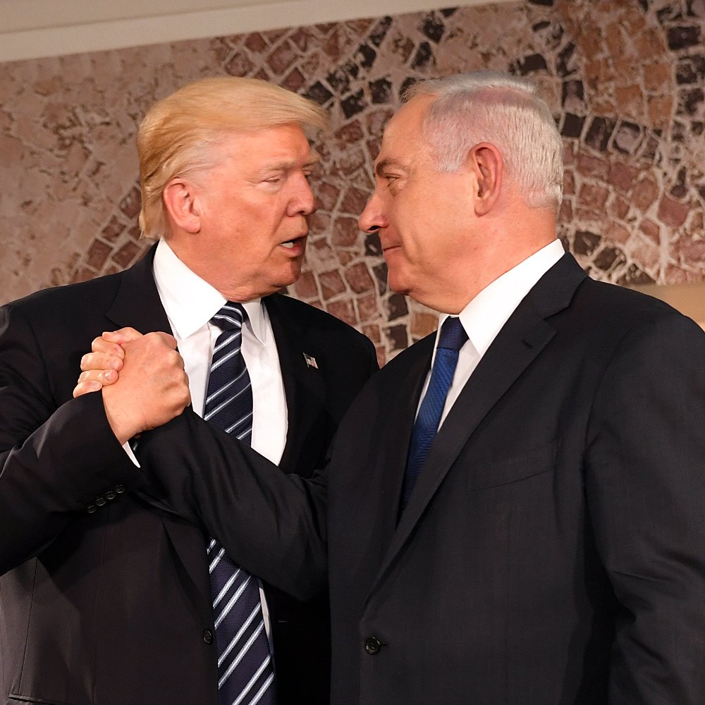 L'image montre deux hommes en train de se serrer la main avec des expressions sérieuses. Ils sont habillés en costume, l'un portant une cravate rayée et l'autre une cravate unie. L'arrière-plan possède des motifs décoratifs. Cette scène semble se dérouler lors d'un événement officiel ou d'une rencontre diplomatique.