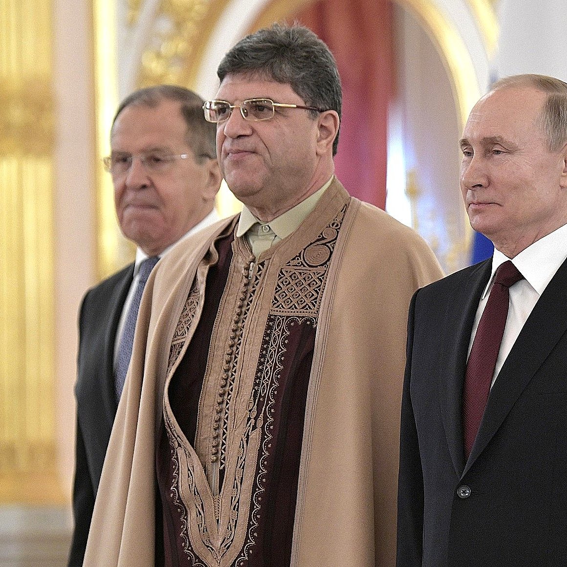 La imagen muestra a tres hombres de pie en un entorno ceremonial. El hombre a la izquierda lleva un traje oscuro y tiene una expresión seria. En el centro, el segundo hombre viste una prenda con un diseño decorativo y también parece estar en una postura formal. A la derecha, se encuentra otro hombre, vestido con un traje oscuro y corbata, que observa al frente. El fondo tiene un ambiente lujoso con detalles dorados y alusiones a un contexto oficial.