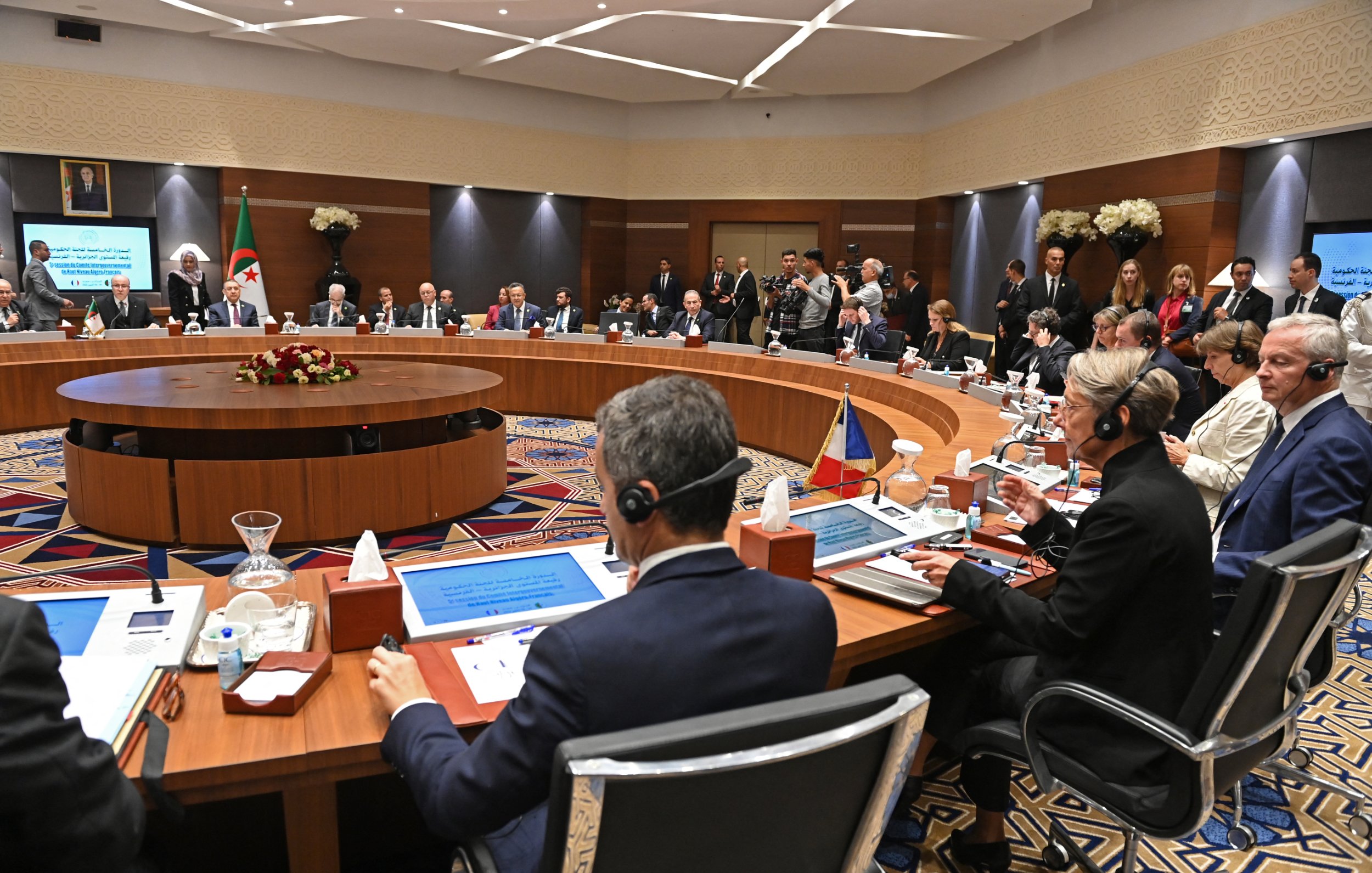 L'image montre une réunion formelle dans une salle de conférence. De nombreuses personnes sont assises autour d'une grande table ovale, probablement des représentants de différents pays ou organisations. Certains participants portent des casques d'écoute, indiquant qu'il pourrait y avoir des traductions simultanées. La salle est décorée avec des fleurs et des éléments modernes, et il y a des écrans affichant des informations pertinentes. Certaines personnes se tiennent debout en arrière-plan, observant les discussions. L'atmosphère semble professionnelle et concentrée.