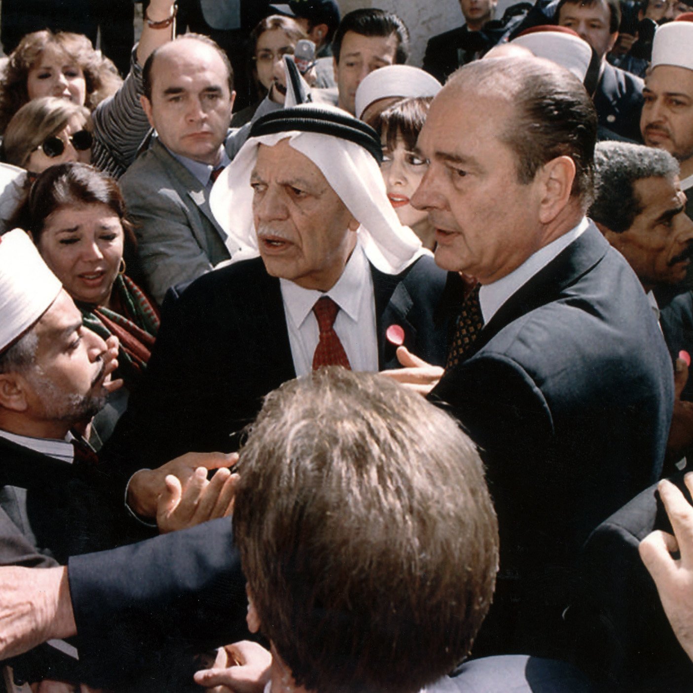 L'image montre une scène chargée d'émotion et de tension. On y voit plusieurs personnes rassemblées autour d'une figure centrale, visiblement un homme en costume. D'autres personnes, portant des vêtements traditionnels et des turbans, semblent interagir avec lui. L'atmosphère est dynamique, avec des visages concentrés et des gestes animés, ce qui suggère une discussion ou un événement important. Le mélange de styles vestimentaires indique une diversité culturelle parmi les personnes présentes, ce qui pourrait refléter un moment significatif sur le plan social ou politique.