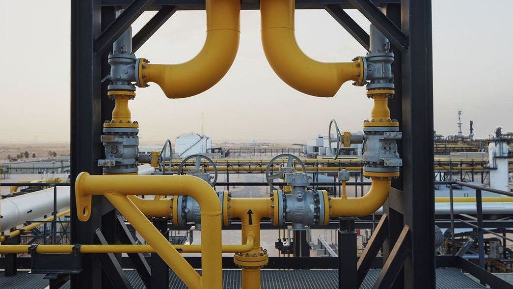 The image features a complex system of industrial piping. Prominent yellow pipes are connected at various angles, showcasing a network of valves and fittings. The background reveals an industrial landscape, with large storage tanks and equipment visible. The overall scene conveys a sense of heavy machinery and infrastructure typically found in oil or gas facilities. The sky appears clear, indicating it might be a time of day either in the morning or late afternoon.