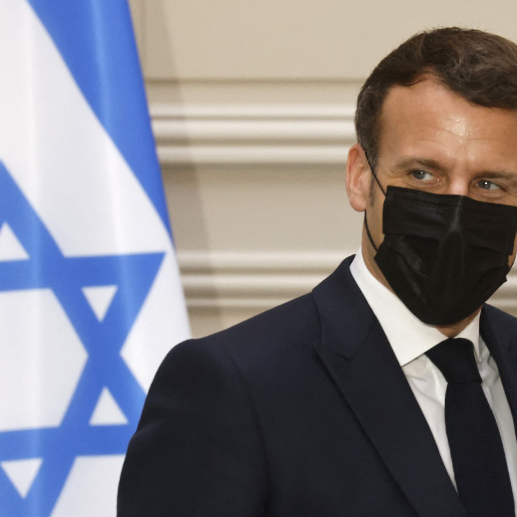The image shows a man in a dark suit wearing a black face mask. He appears to be looking slightly to the side, with a serious expression. In the background, there is a flag featuring a blue Star of David on a white field, which likely signifies that the setting is related to Israel. The overall atmosphere suggests a formal or diplomatic context.