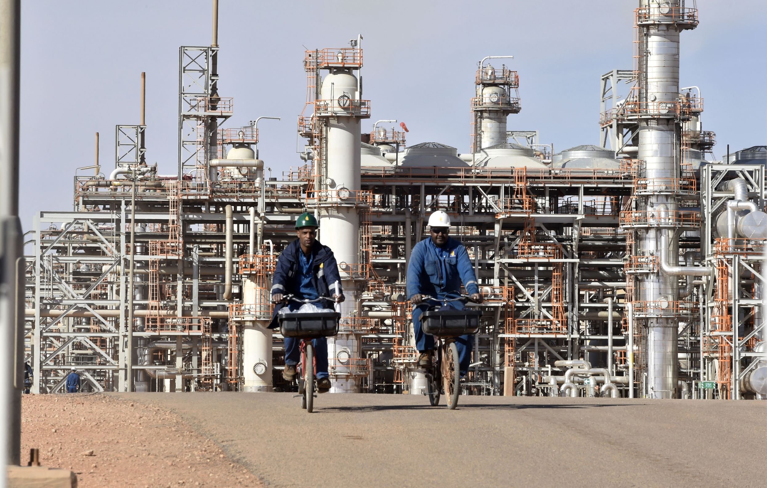 L'image montre deux hommes à vélo, portant des vêtements de travail et des casques, qui circulent sur une route. En arrière-plan, on peut voir une installation industrielle avec de grandes structures métalliques et des pipelines, typiques d'une raffinerie ou d'un complexe pétrochimique. Le ciel est clair et les installations semblent actives, témoignant d'un environnement de travail industriel.