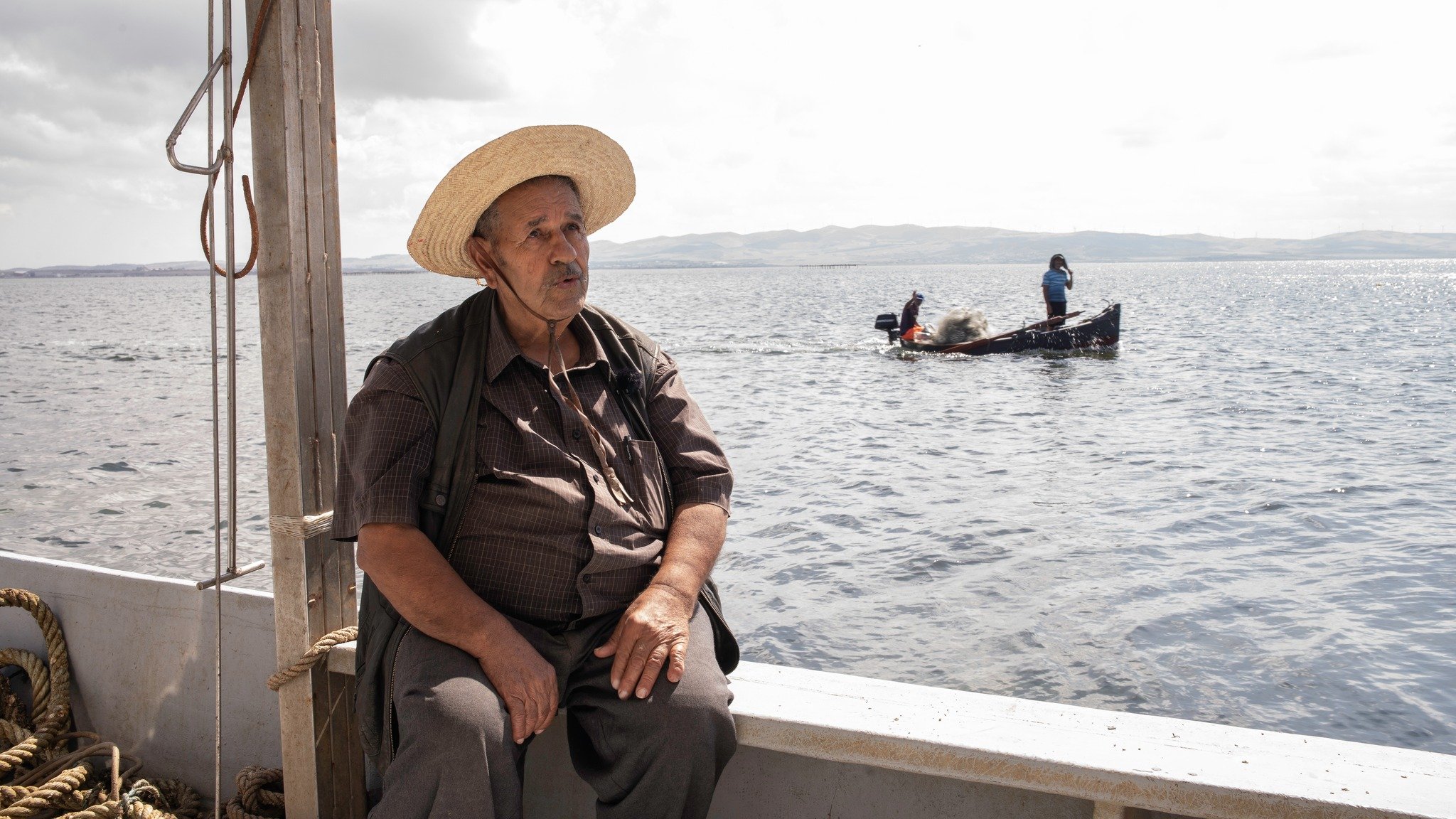 L'image montre un homme assis sur le bord d'un bateau, portant un chapeau de paille large. Il a l'air pensif et regarde vers le large. En arrière-plan, on aperçoit un autre bateau avec des personnes qui semblent s'affairer à la pêche. L'environnement est calme, avec des eaux calmes et un ciel légèrement nuageux, suggérant une ambiance paisible.