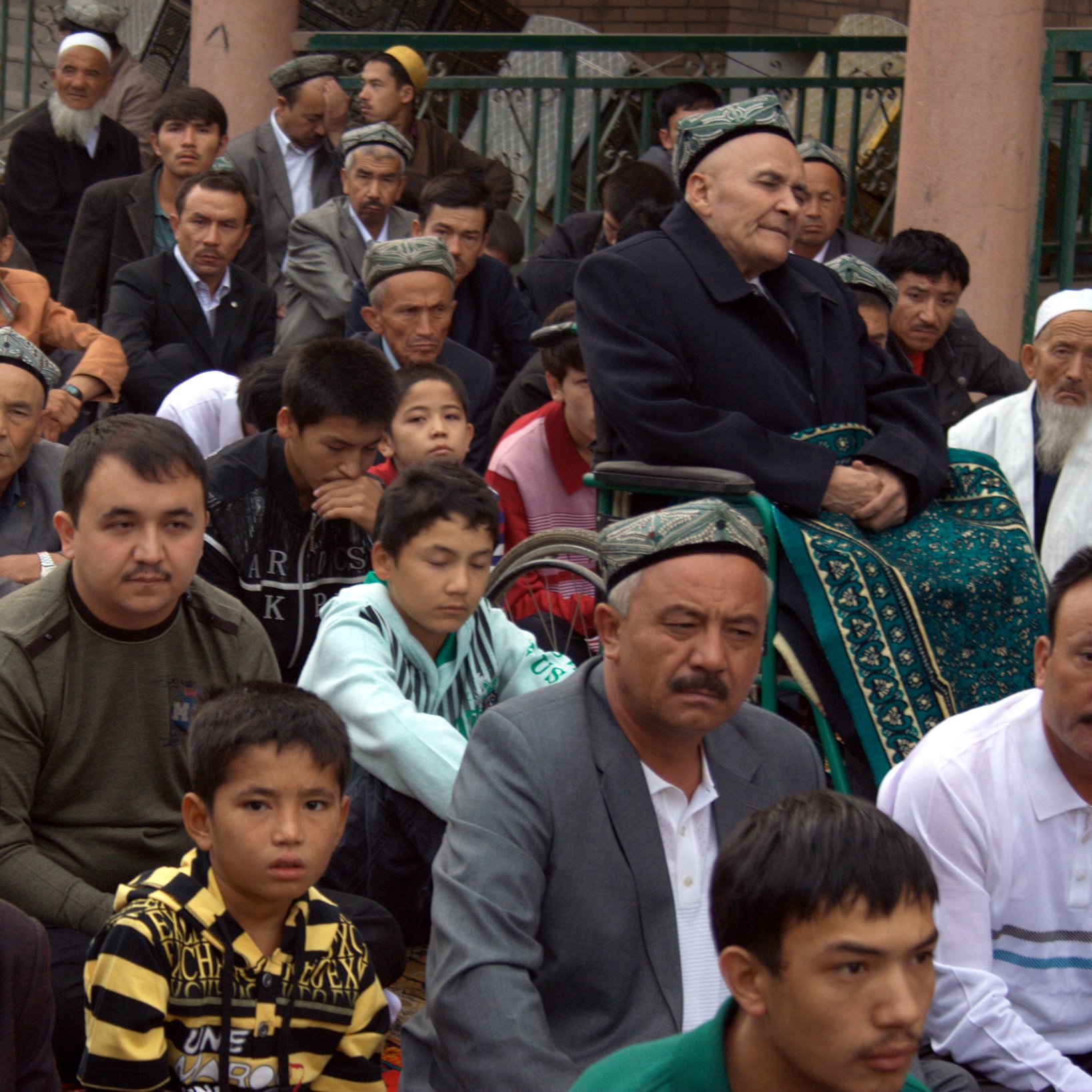The image depicts a group of individuals seated together, primarily men, in a communal setting. They appear to be engaged in a gathering or possibly a religious ceremony, with some wearing traditional attire. In the foreground, there is an elderly man seated in a wheelchair, and others are attentively looking forward. The overall atmosphere reflects a sense of community and shared purpose. The individuals show a range of ages, from children to older men, indicating that the event likely holds significance for various generations.