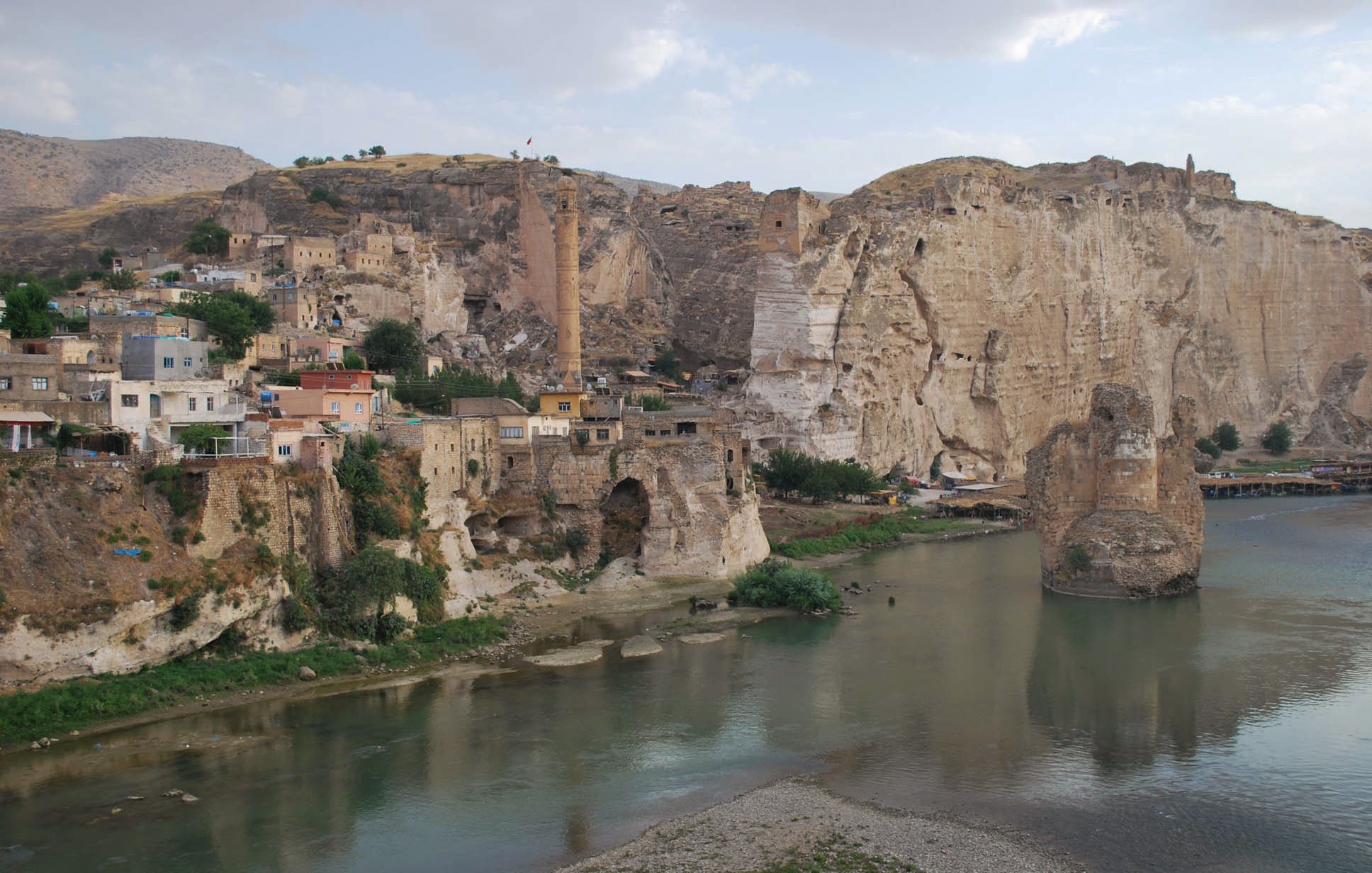 The image depicts a scenic landscape featuring a river flowing through a valley. On the left side, there are rocky cliffs and remnants of ancient structures, hinting at a historical settlement. The area appears to be a mix of natural and man-made elements, with a few buildings clinging to the hillside. In the background, there are more cliffs and a prominent tower-like structure, suggesting a rich history and possibly a fortified site. The sky is partly cloudy, adding to the serene atmosphere of the scene.