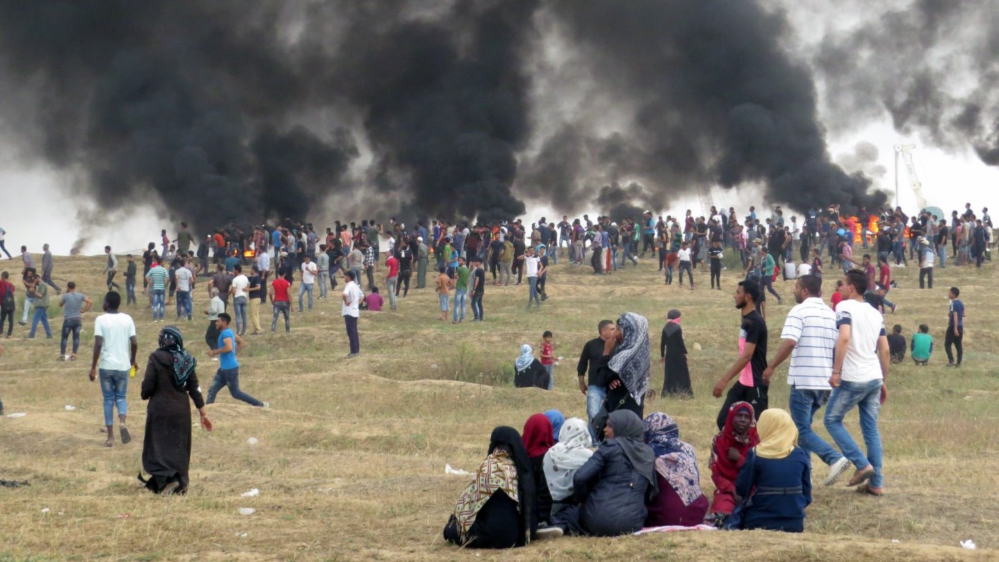 L'image montre une grande foule de personnes rassemblées sur une colline, avec d'importants nuages de fumée noire émanant en arrière-plan. Les gens semblent se tenir debout ou marcher, certains regardant vers la fumée. Au premier plan, un groupe de personnes est assis sur le sol, tandis que d'autres se déplacent autour d'eux. L'ambiance est tendue, indiquant une situation d'agitation ou de protestation. Le paysage est principalement sec avec quelques herbes.
