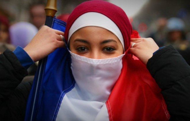 The image features a young woman dressed in a red and white outfit, with a hijab, who is adjusting her clothing. She is holding a flag that represents France, with the colors of blue, white, and red. The background suggests a public gathering or demonstration, with people present in a city setting. The atmosphere appears to convey themes of identity and cultural expression.
