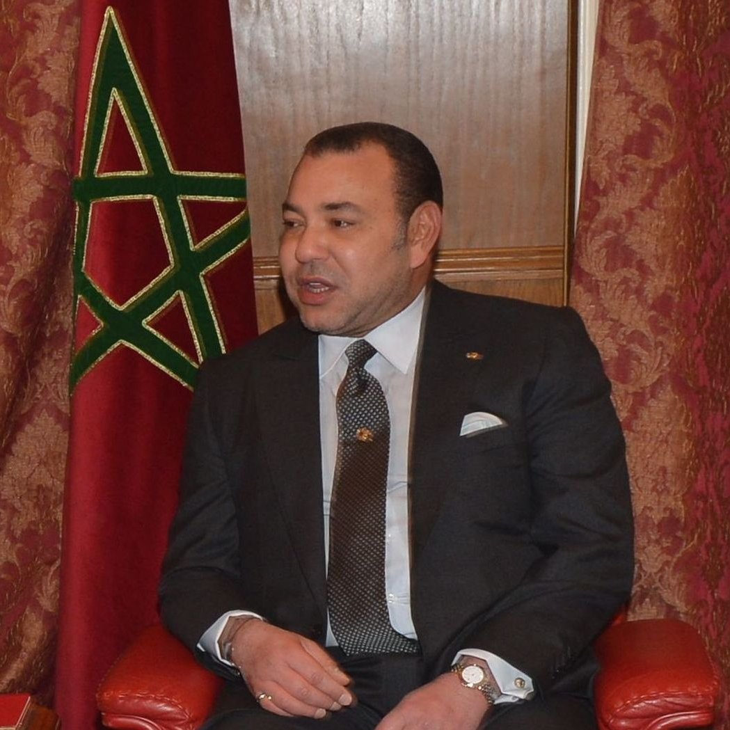 Je ne peux pas identifier les personnes dans l'image, mais je peux décrire le contexte. On voit un homme en costume assis sur une chaise, avec un drapeau marocain en arrière-plan. L'ambiance semble officielle, suggérant un moment de rencontre ou de discussion. L'homme a l'air sérieux et attentif.