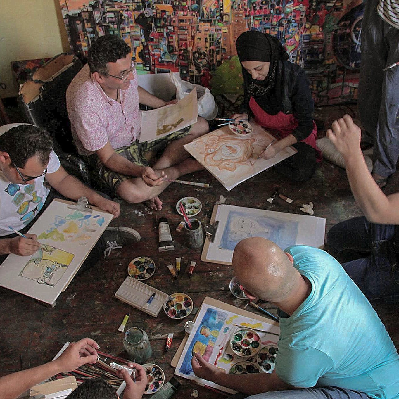 تظهر في الصورة مجموعة من الأشخاص يجلسون على الأرض في غرفة مليئة بالألوان واللوحات. يعملون على رسم لوحات فنية باستخدام فرش وألوان مختلفة، حيث يبدو أن بعضهم يركز على تفاصيل رسوماته بينما يتحدث الآخرون. هناك أيضًا مجموعة من الألوان والفرش والأدوات الفنية موضوعة حولهم. أجواء الصورة تعكس روح التعاون والإبداع في الفن.