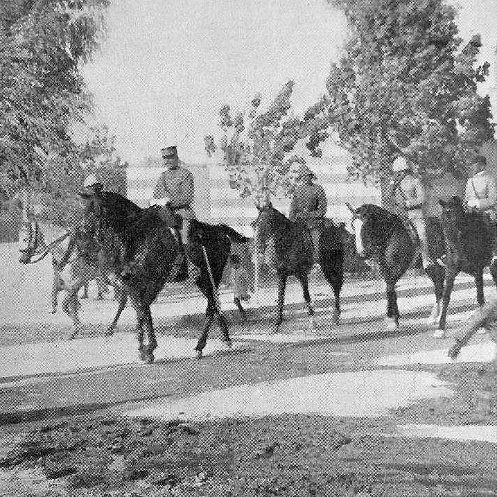 در این تصویر، تعدادی سوارکار باuniform نظامی در حال عبور از یک خیابان هستند. آنها بر روی اسب‌های خود سوارند و به سمت جلو حرکت می‌کنند. در کنار خیابان، یک مرد پیاده در حال عبور است و به سمت دیگر می‌رود. در پس‌زمینه، ساختمان‌هایی دیده می‌شوند که شاید مربوط به دوره تاریخی خاصی باشند و همچنین یک مناره یا برج در فاصله‌ای دور قرار دارد. درختان اطراف نیز به تصویر حال و هوای زمان قدیم را می‌دهند.