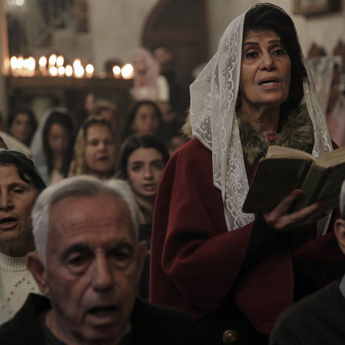 در این تصویر، تعدادی از افراد در یک کلیسا یا مکان مذهبی جمع شده‌اند. یک زن با پوشش خاص و حجابی زیبا در حال خواندن یک کتاب مقدس به نظر می‌آید. او در کانون توجه قرار دارد و دیگر افراد به او نگاه می‌کنند یا در حال خواندن دعا هستند. جو کلی تصویر معنوی و نسبتاً احساسی به نظر می‌رسد، با نور ملایمی که از شمع‌ها و فضای داخلی کلیسا می‌آید.