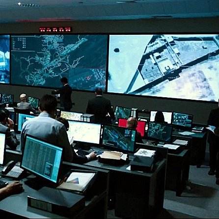 L'image montre une salle de commandes avec de nombreuses personnes travaillant devant des ordinateurs. Il y a plusieurs écrans géants affichant des cartes et des informations, et l'atmosphère semble intense et concentrée. Les employés, habillés en costumes ou en tenues professionnelles, semblent analyser des données ou surveiller des opérations en temps réel. La pièce est éclairée par la lumière des écrans, créant une ambiance technologique et dynamique.