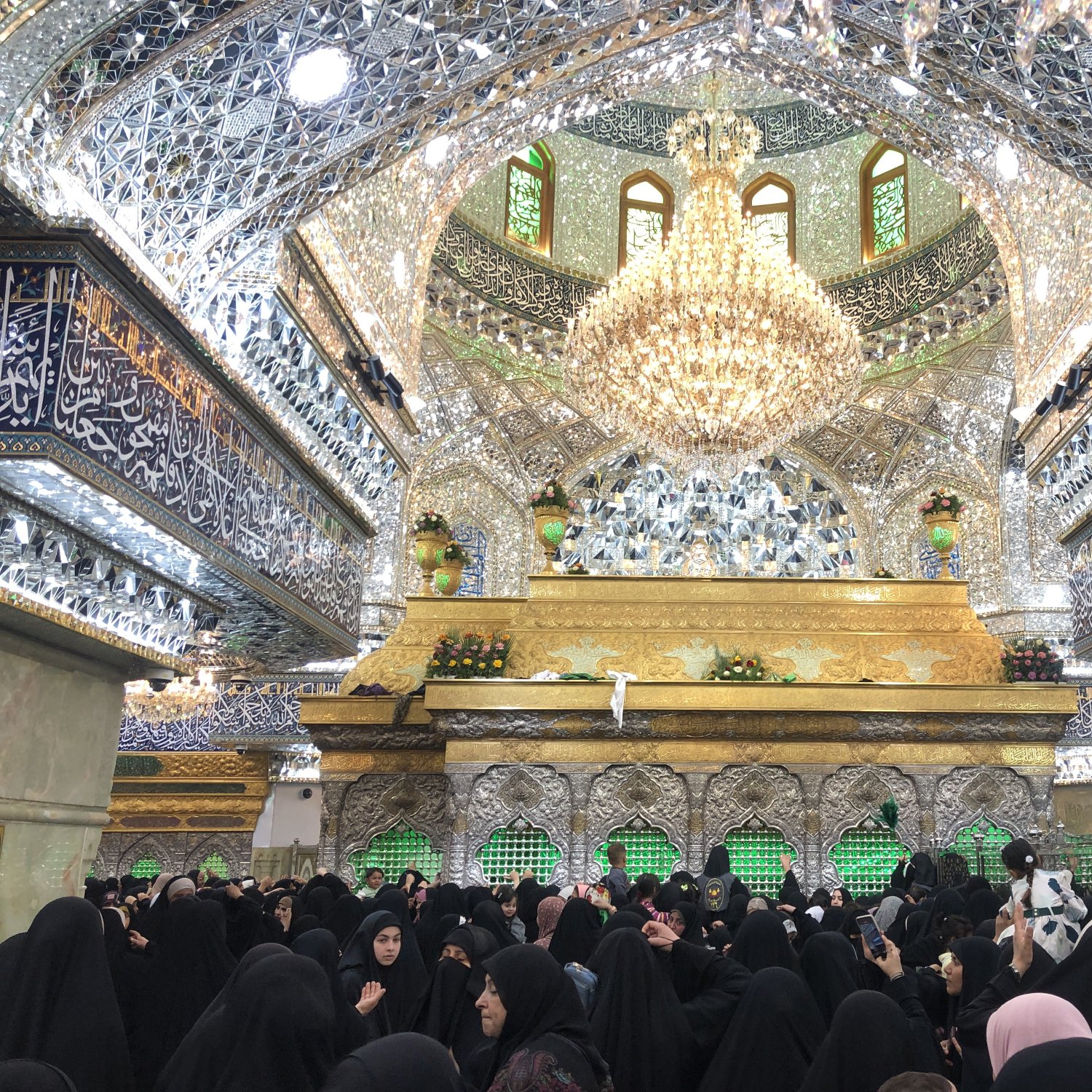 الصورة تظهر مشهداً داخلياً في مكان ديني، حيث تتواجد الكثير من النساء المحجبات داخل قاعة مزينة بتفاصيل معمارية جميلة ومزيّنة بالزخارف والجدران المغطاة بالزجاج اللامع. يتوسط القاعة مصباح ضخم معلق، ويبدو أن هناك ضريحاً أو مكان مقدس في الخلفية مزين بالعناصر الذهبية والكتابات الإسلامية. الأجواء تعكس روحانيّة وعمق الإيمان، حيث يتجمع الناس في مكان يتسم بالهدوء والتأمل.