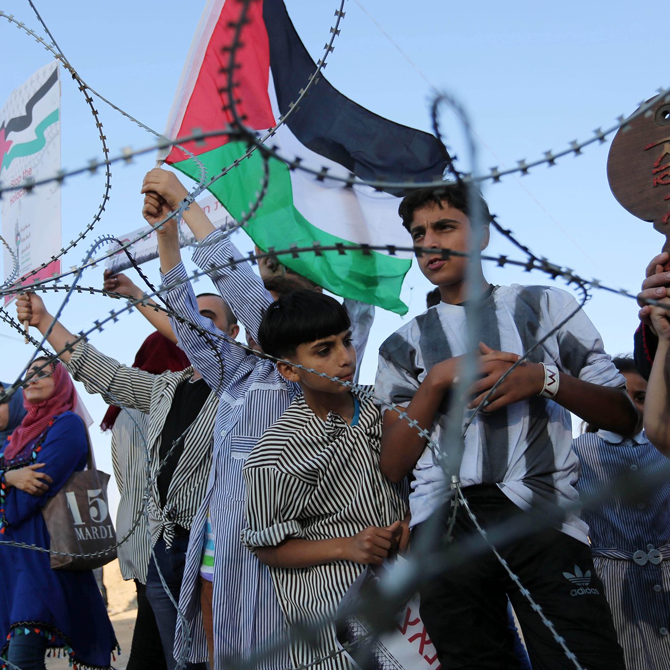 در این تصویر، گروهی از مردم، از جمله کودکانی، در نزدیکی یک نوار سیم‌خاردار تجمع کرده‌اند. برخی از آن‌ها پرچم فلسطین را در دست دارند و به نظر می‌رسد که در حال ابراز احساسات یا مخالفت با شرایطی هستند. لباس‌های مختلفی بر تن دارند و حالت‌هایشان متنوع است. زمینه تصویر آسمانی صاف و روشن به نظر می‌رسد.