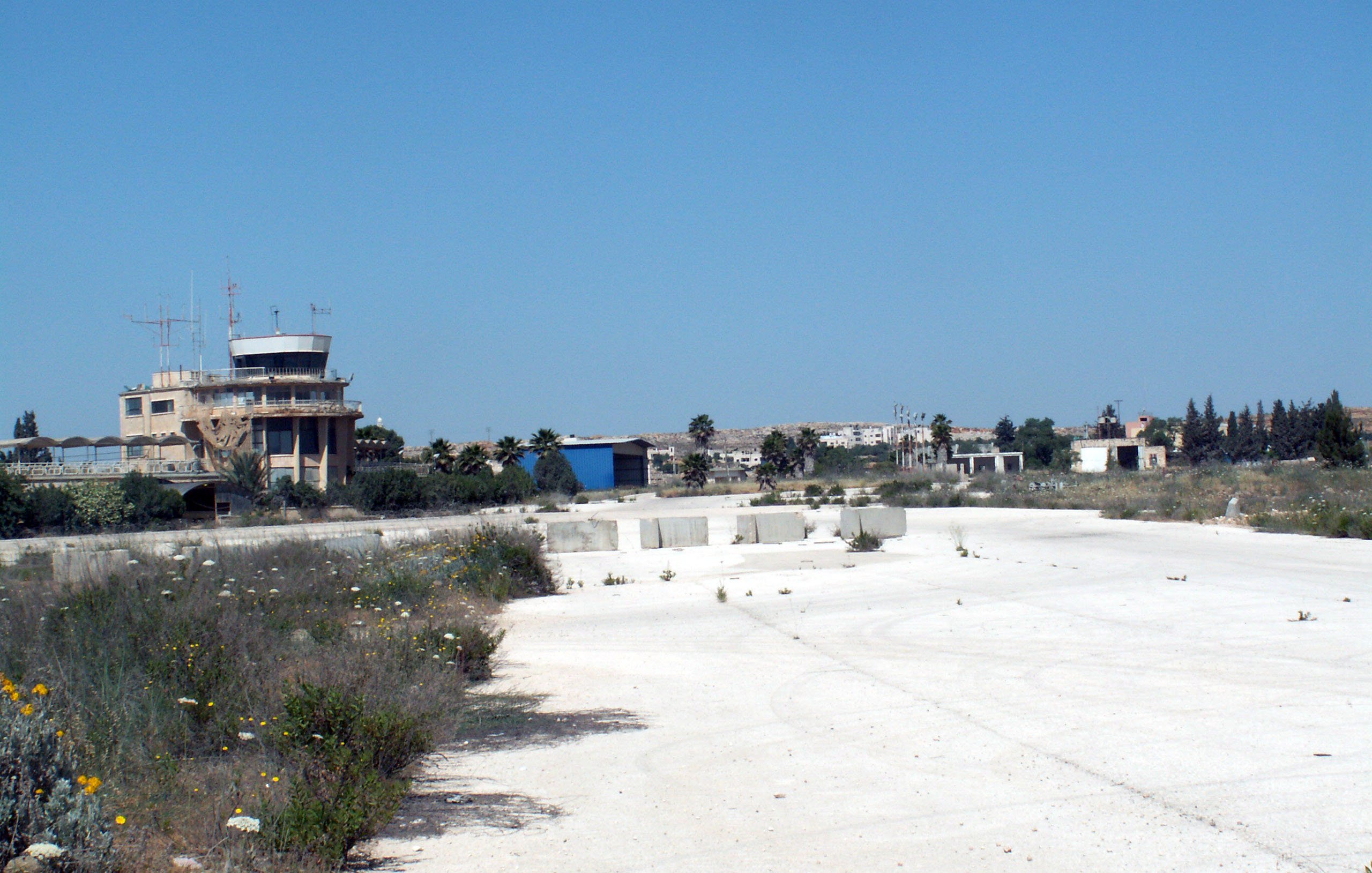 The image depicts an abandoned or derelict area, likely an old airstrip or airport. In the background, there is a control tower or building, which appears to be in disrepair. Surrounding the area are sparse patches of vegetation, including some wildflowers, and the ground is mostly composed of white gravel or concrete. The sky is clear and blue, indicating a bright, sunny day, and the overall scene conveys an atmosphere of neglect and desolation.