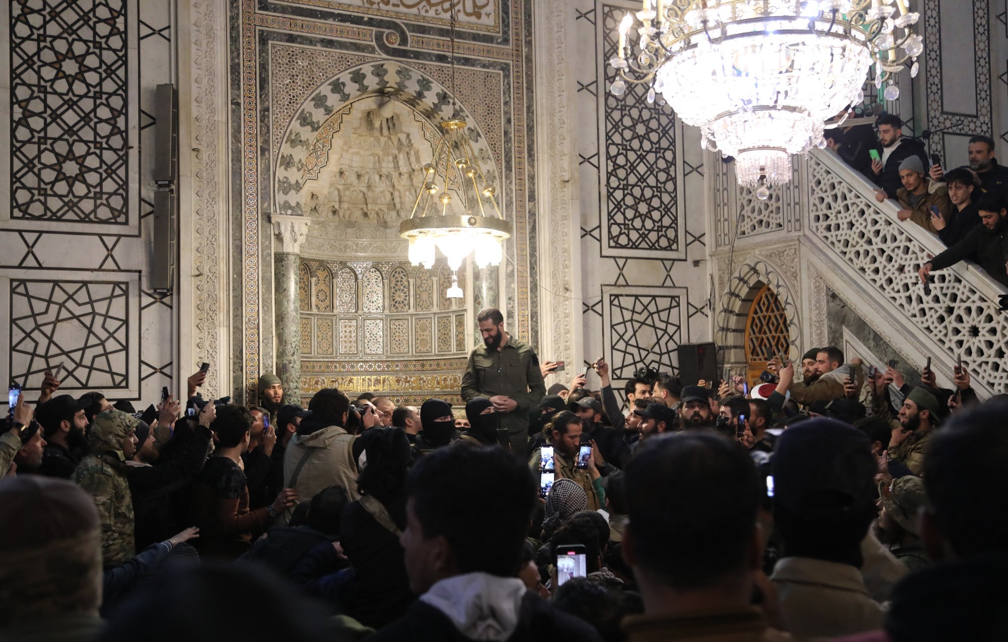 L'image montre un intérieur richement décoré, probablement d'un bâtiment historique ou religieux, avec des motifs géométriques et des ornements élaborés. Au centre, une personne se tient en hauteur, entourée d'une foule qui utilise des téléphones pour capturer le moment. Des lumières brillantes, notamment un lustre, illuminent la scène. L'atmosphère semble être celle d'un rassemblement ou d'un événement significatif.