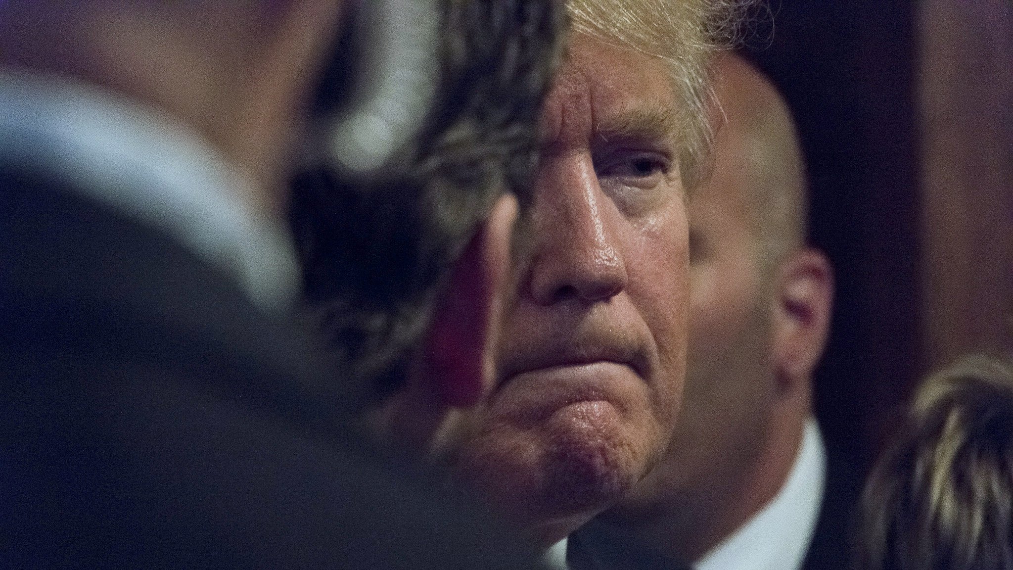 در این تصویر، یک مرد با موهای روشن و کمی حالت خمیده سر دیده می‌شود. او در حالتی contemplative به نظر می‌رسد و گونه‌ها و خطوط صورتش مشخص است. در اطراف او چندین فرد دیگر مشاهده می‌شوند که به نظر می‌رسد در حال گفتگو یا مراقبت از او هستند. پس‌زمینه تصویر تاریک و مبهم است، که تمرکز را بر روی چهره او بیشتر می‌کند. این تصویر احساس جدیت و تفکر عمیق را منتقل می‌کند.