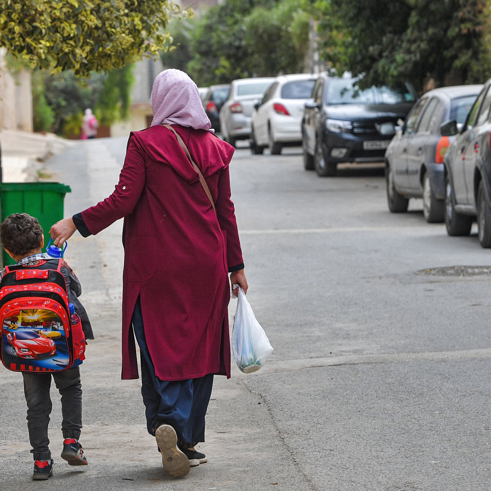 The image shows a woman walking down a street with a young child. The woman is wearing a long maroon coat and a headscarf, while the child has a colorful backpack with a cartoon character design. They are walking hand in hand along a road lined with parked cars and greenery on the sides. The atmosphere appears casual and everyday, suggesting they are possibly returning from school or heading to a local destination.
