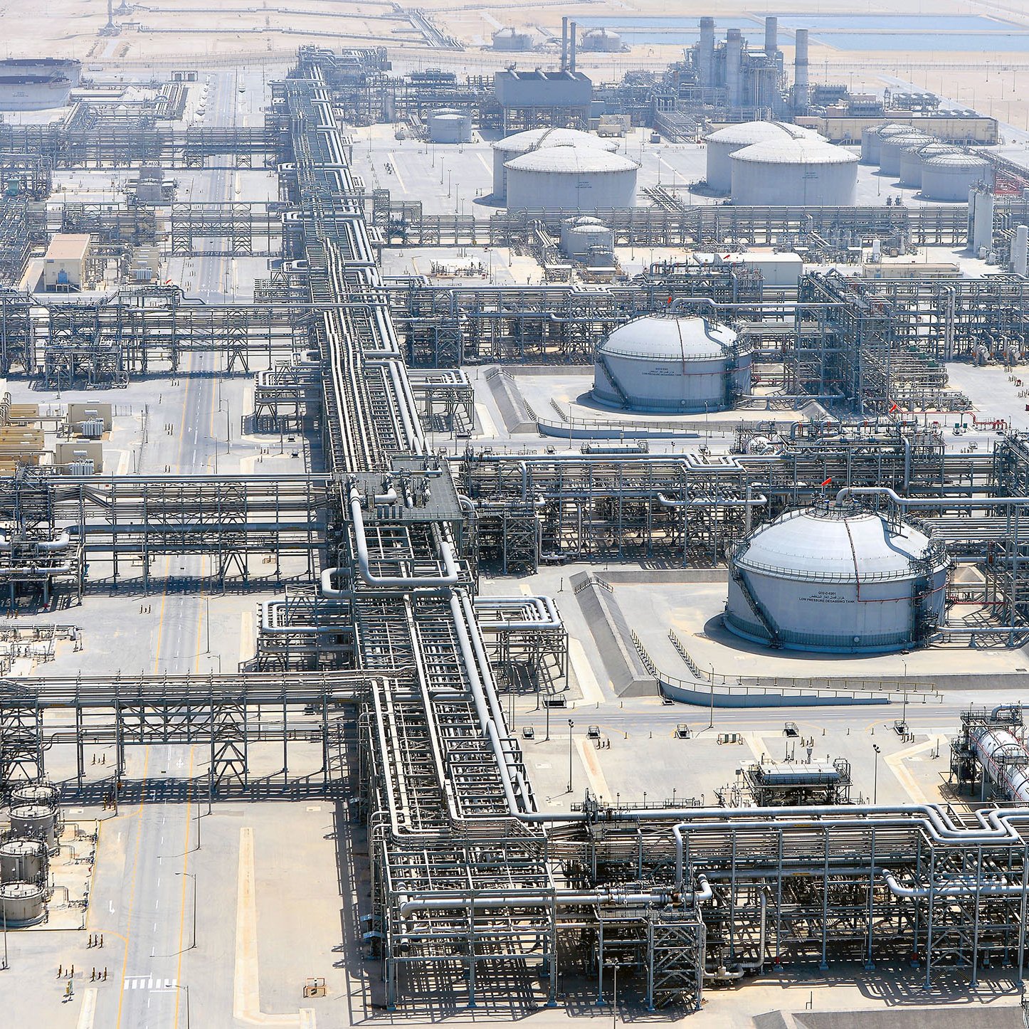 La imagen muestra una vista aérea de una planta industrial, probablemente relacionada con la producción o el refinado de petróleo. Se pueden observar grandes tanques de almacenamiento, tuberías extensas que se entrelazan por todo el área y estructuras de soporte. El entorno es desértico, con un paisaje árido al fondo, lo que sugiere que esta instalación podría estar ubicada en una región donde la extracción de recursos naturales es común. La organización y complejidad de las tuberías y tanques reflejan la sofisticación de la infraestructura industrial.