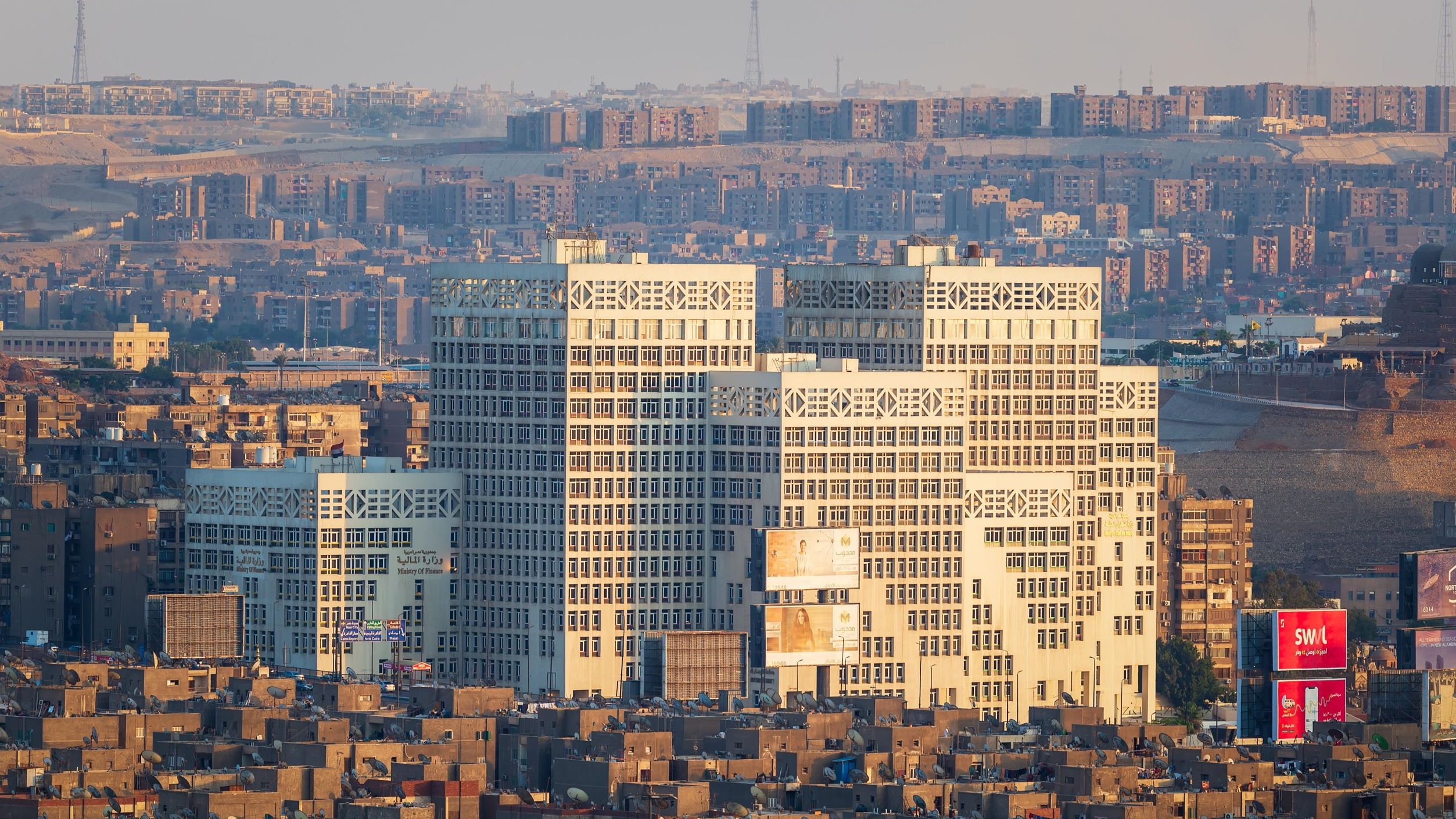 The image depicts a cityscape featuring a cluster of modern buildings in the foreground, characterized by their distinct architectural design with large glass windows. These buildings stand out against a backdrop of densely packed smaller structures in varying shades of brown. The scene conveys a contrast between contemporary architecture and older, more traditional urban forms. The setting appears to be at a distance, possibly showing a skyline during early morning or late afternoon when the light casts warm tones on the buildings. The surrounding area is marked by hilly terrain and a sprawling development, illustrating a bustling urban environment.