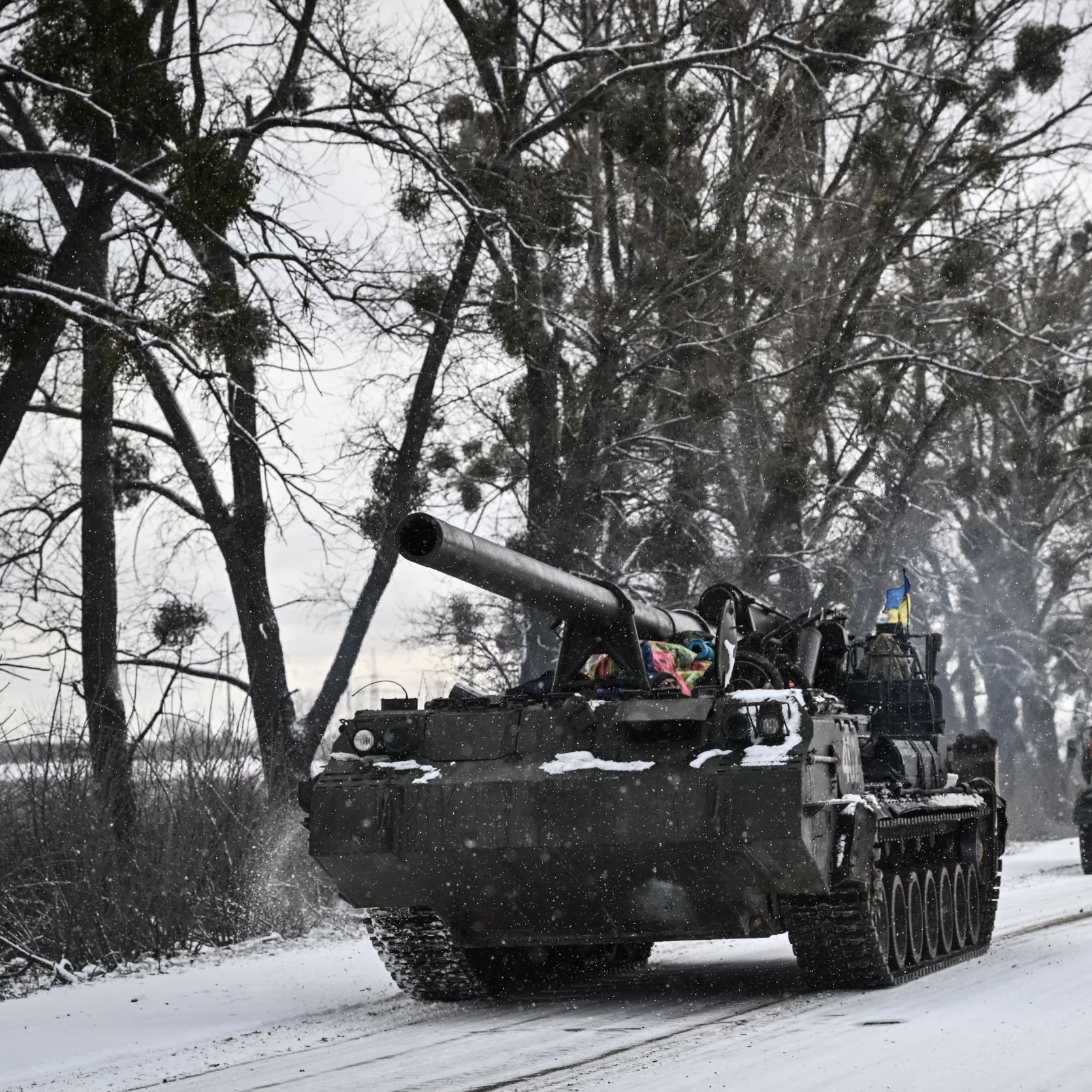 L'immagine mostra un carro armato che si muove lungo una strada innevata, circondato da alberi. Il carro armato ha una canna protrudente e presenta dettagli decorativi che potrebbero indicare l'appartenenza a una specifica unità militare. Accanto al carro armato, si intravedono anche alcuni veicoli, probabilmente militari, che viaggiano sulla stessa strada. Il paesaggio è caratterizzato da un'atmosfera invernale, con la neve che copre sia la strada che il terreno circostante.