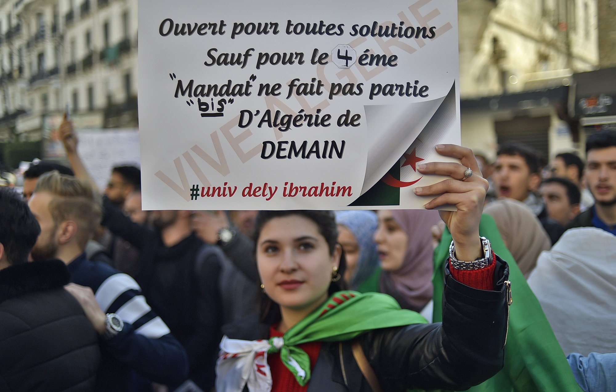 L'image montre une manifestation, où une jeune femme tient une pancarte. La pancarte indique qu'elle est ouverte à toutes solutions, sauf pour le 4ème mandat, affirmant que le mandat ne fait pas partie de l'Algérie de demain. La femme est entourée d'autres manifestants et porte un drapeau. L'atmosphère semble engagée et déterminée, reflétant des revendications politiques.