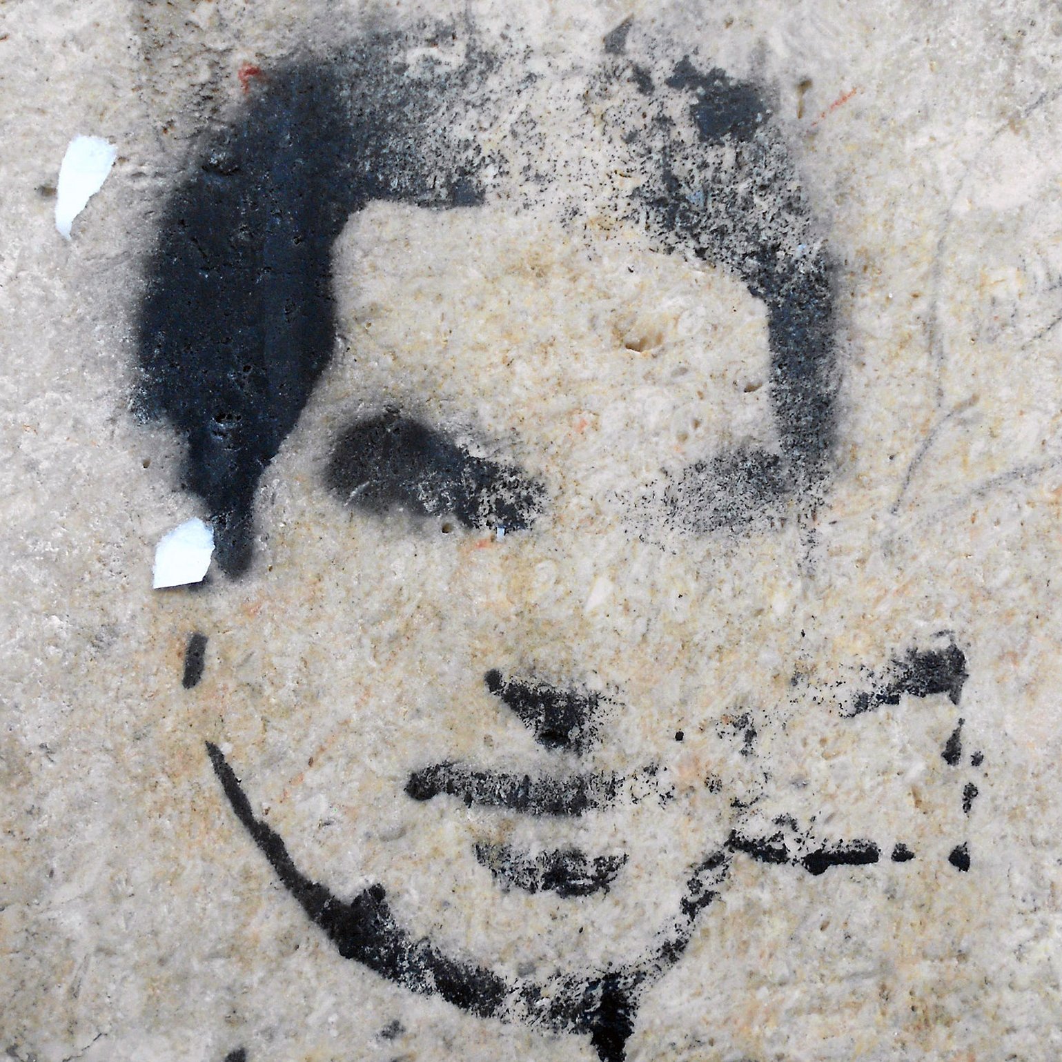 الصورة تظهر رسومًا جدارية تُظهر وجه إنسان بأسلوب الطلاء الرش. يبدو أن الرسم مقطوع بعض الشيء، مع تفاصيل غامضة لفم وأنف وعينين. الخلفية غير منتظمة وقد تشتمل على بعض آثار الندوب أو التآكل، مما يعطي الانطباع بأنها قد رسمت على سطح قديم أو متآكل. اللون المستخدم في الرسمة أسود بينما الخلفية ذات لون فاتح.