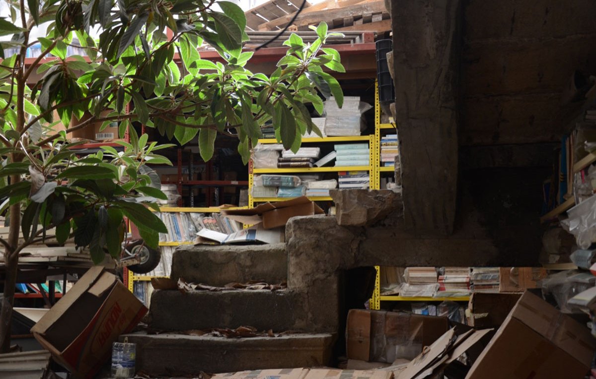 الصورة تظهر مشهدًا داخليًا لمكان يبدو وكأنه مستودع أو مكتبة مهجورة. يوجد في الصورة درج متآكل يقود إلى مستوى أعلى، محاط بكومة من الصناديق والكتب الملقاة على الأرض. على اليمين، هناك أشجار أو نباتات خضراء تنمو في المكان، مما يعطي لمسة طبيعية للمشهد. في الخلفية، تتواجد رفوف مزدحمة بالكتب والصناديق. الإضاءة تأتي من النوافذ العليا، مما يضيف عمقًا وإحساسًا بالغموض للمكان.