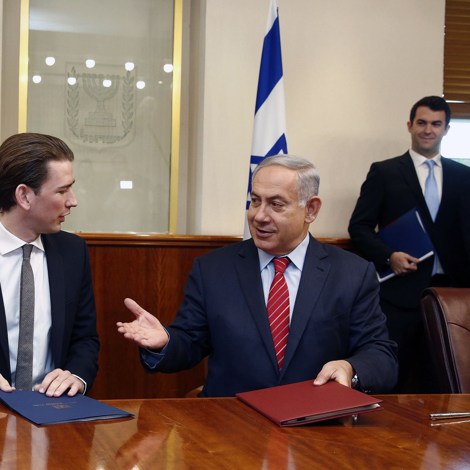 در این تصویر، دو مرد در حال گفتگو و گفت‌وگو هستند. یکی از آنها در سمت چپ، کت و شلوار تیره و کروات دارد و ظاهراً جوانتر به نظر می‌رسد. مرد دیگر در سمت راست، کت و شلوار رسمی به تن دارد و با حرکات دست خود در حال بیان مطلبی است. در پس‌زمینه، پرچم‌هایی به چشم می‌خورند و فضایی رسمی به نظر می‌رسد. نمای کلی تصویر نشان‌دهنده یک جلسه یا توافق رسمی است.