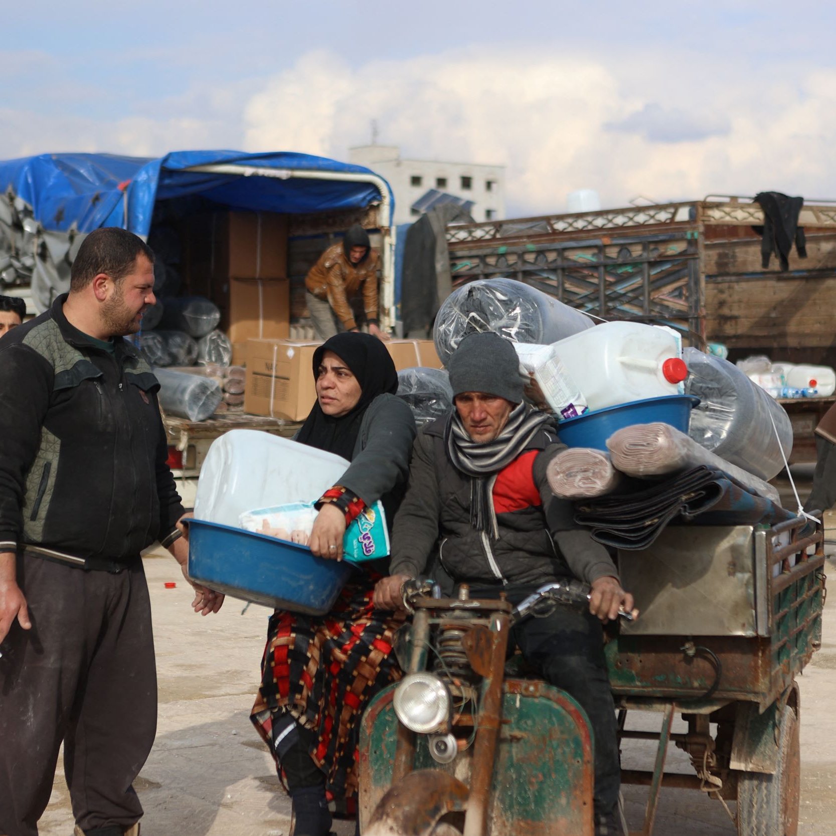 الصورة تظهر تجمعاً في منطقة يبدو أنها مكان لتوزيع المساعدات. يوجد رجل وامرأة يحملان بعض الأغراض، بينما يجلس رجل آخر على عربة صغيرة تحمل زجاجات ومحتويات أخرى. تتواجد خلفهم مجموعة من الأشخاص الذين يعملون على تحميل وتفريغ الأغراض من الشاحنات. السماء تبدو ملبدة بالغيوم، مما يعطي انطباعًا عن الأجواء المحيطة.