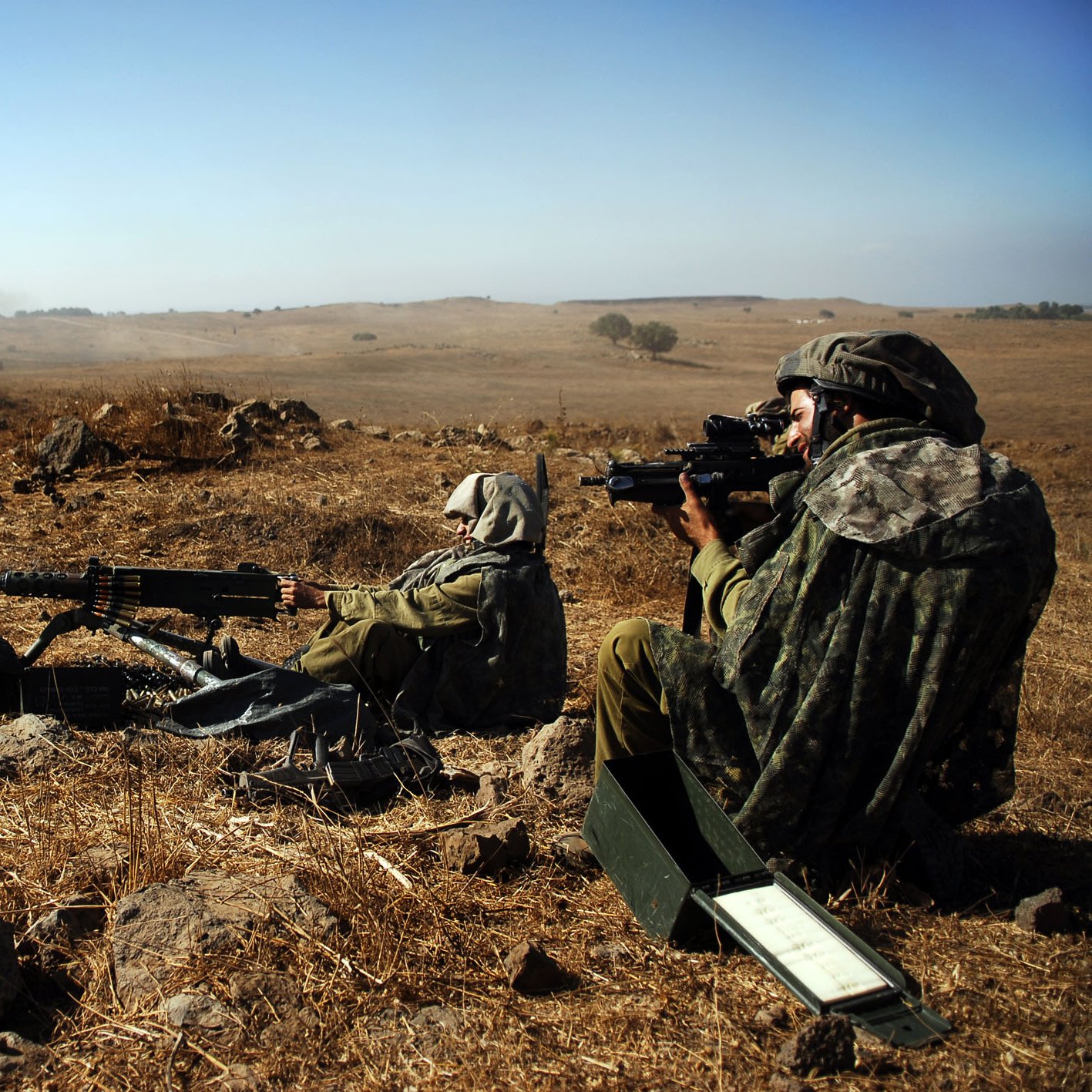 Sur l'image, on peut voir deux soldats en position de tir dans un paysage aride. Ils sont vêtus de tenue de camouflage et portent des casques. L'un des soldats est à genoux, prêt à tirer avec une mitrailleuse, tandis que l'autre est assis, observant à travers un dispositif de visée. Le terrain est composé de rochers et de terre sèche, et le ciel est dégagé, indiquant une journée ensoleillée. Cette scène évoque un environnement militaire austère et une préparation à l'action.