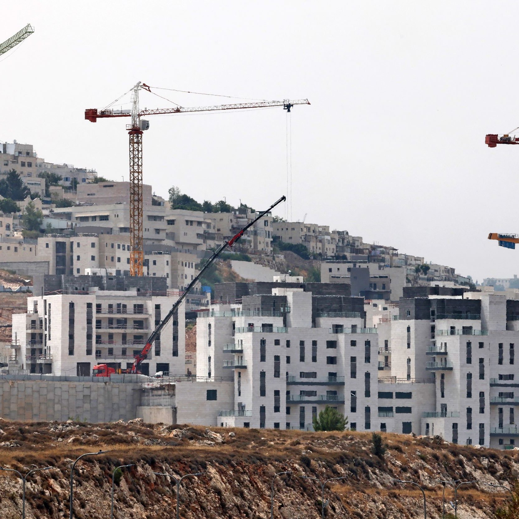 L'image montre un paysage urbain en construction. On peut voir plusieurs grues de construction sur des bâtiments en cours d'érection. Les structures, encore inachevées, présentent des façades en béton. À l'arrière-plan, on aperçoit des collines et d'autres constructions, indiquant un développement résidentiel dans la région. Le ciel semble brumeux, ce qui donne une ambiance un peu sombre à la scène.