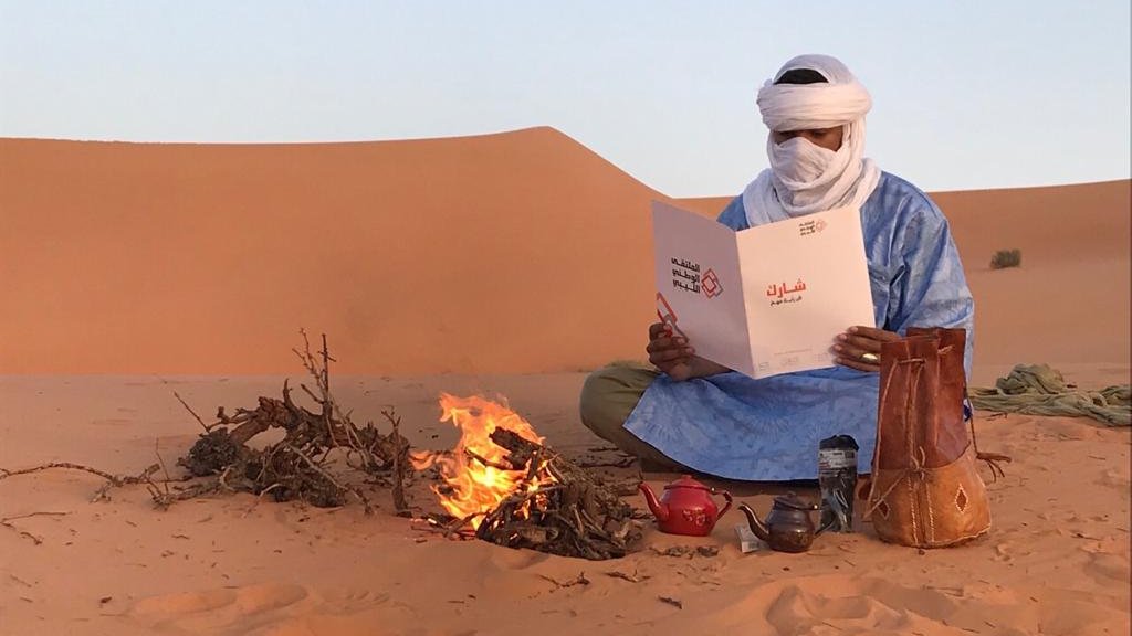 L'image montre un homme assis dans le désert, portant des vêtements traditionnels bleus et un turban. Il est devant un petit feu de camp, où des brindilles brûlent. L'homme tient un menu qu'il examine attentivement. À côté de lui, il y a une théière et un pot en terre cuite. En arrière-plan, on peut voir des dunes de sable sous un ciel clair. L'ambiance évoque la tranquillité et la culture nomade.