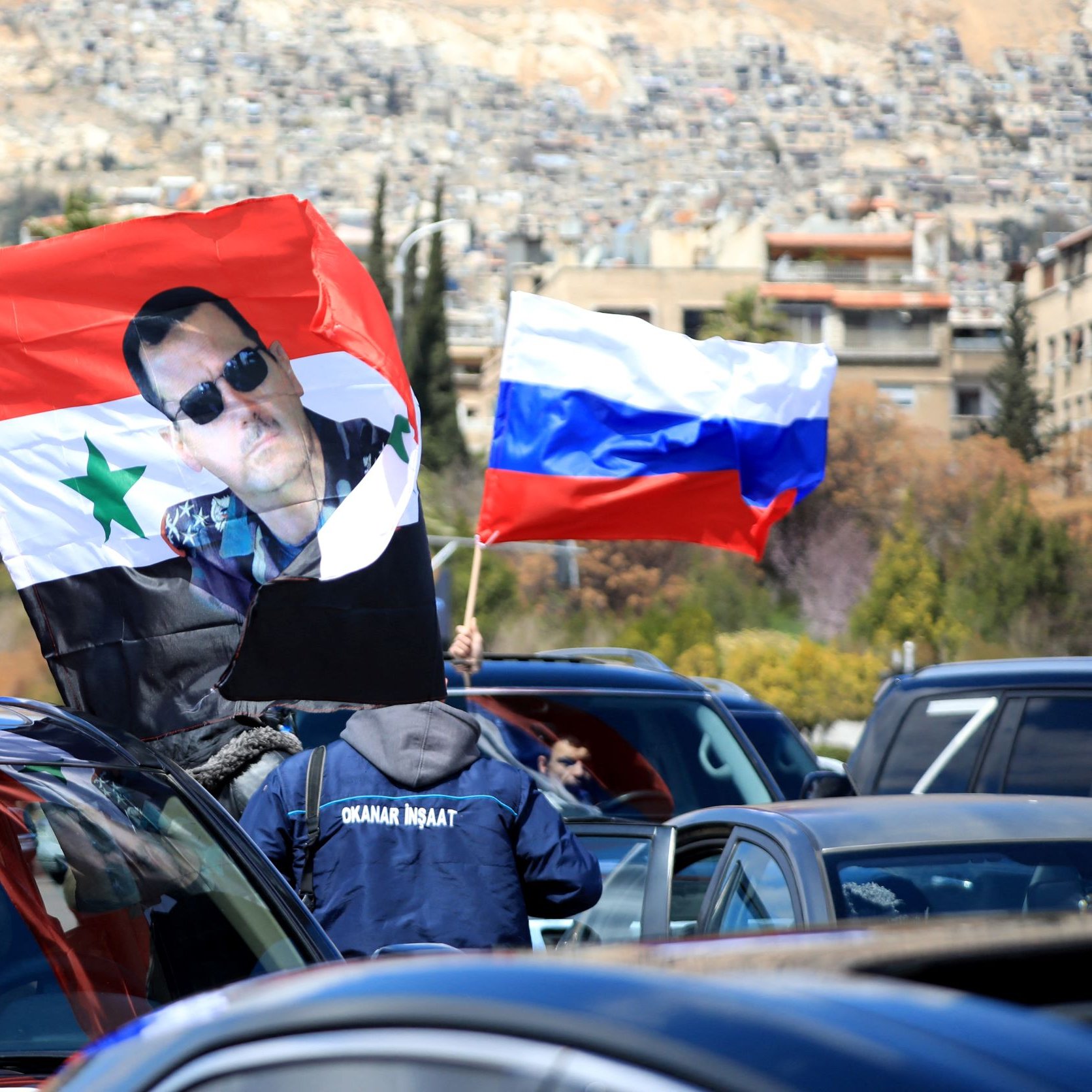 L'immagine mostra una scena di una manifestazione o di una celebrazione in cui sono presenti diverse bandiere. In primo piano si possono vedere bandiere della Russia e una bandiera con un'immagine di un individuo che indossa occhiali da sole e ha un'icona della Siria. Le auto nel contesto suggeriscono che la manifestazione potrebbe avere una certa rilevanza politica o culturale. Sullo sfondo si notano edifici e una zona collinare.