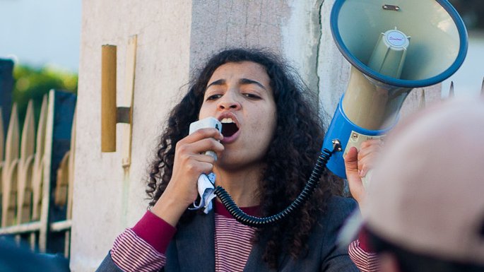 L'image montre une personne debout, utilisant un mégaphone pour s'adresser à un groupe. Elle semble engagée et passionnée, portant un costume avec une chemise à rayures. Son geste et son expression faciale indiquent qu'elle est en train de faire un discours, probablement lors d'une manifestation ou d'un rassemblement. En arrière-plan, des personnes écoutent attentivement, suggérant qu'elle est en train de discuter d'un sujet important. Le cadre général dénote une atmosphère de mobilisation ou d'activisme.