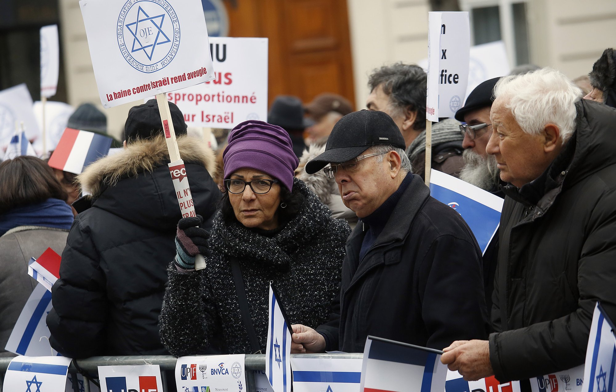 The image depicts a group of protesters gathered in an outdoor setting. They are holding signs and flags, including the Israeli flag and the French flag. The attendees appear to be engaged in a demonstration, showing solidarity or expressing opinions related to Israel. Some individuals are wearing winter clothing, suggesting it is during a colder season. The signs have various messages, possibly related to political or social issues concerning Israel. The atmosphere seems serious, highlighting a significant concern among the participants.