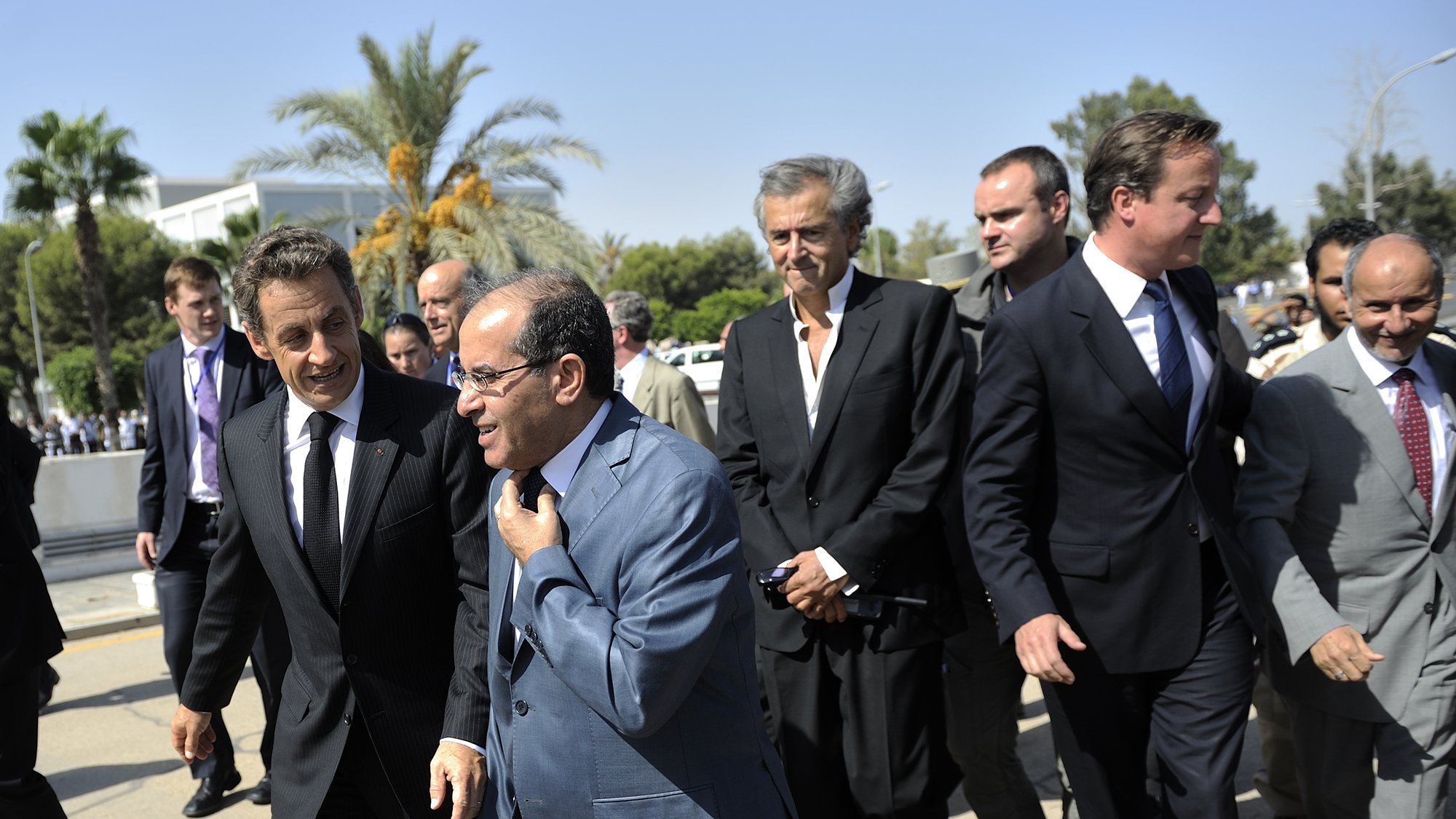 L'image montre un groupe d'hommes marchant ensemble dans un environnement extérieur. Ils semblent être en discussion animée. En arrière-plan, on peut apercevoir des palmiers et un bâtiment, suggérant un climat ensoleillé. Les hommes portent des costumes, ce qui indique un contexte formel ou professionnel. L'ambiance semble dynamique et engageante.