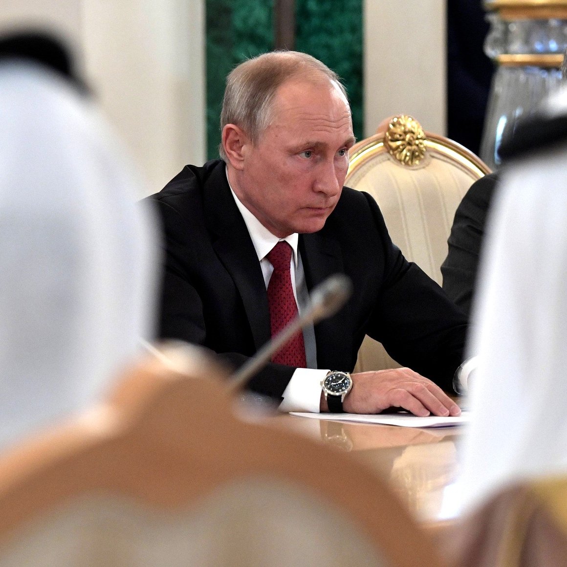 L'image montre un homme assis à une table lors d'une réunion. Il porte un costume noir avec une cravate rouge et semble concentré sur la discussion. Autour de lui, on peut voir des personnes en vêtements traditionnels, comme des robes blanches et des turbans. L'environnement est formel, avec des décorations élégantes. L'atmosphère paraît sérieuse et impliquée dans des délibérations.